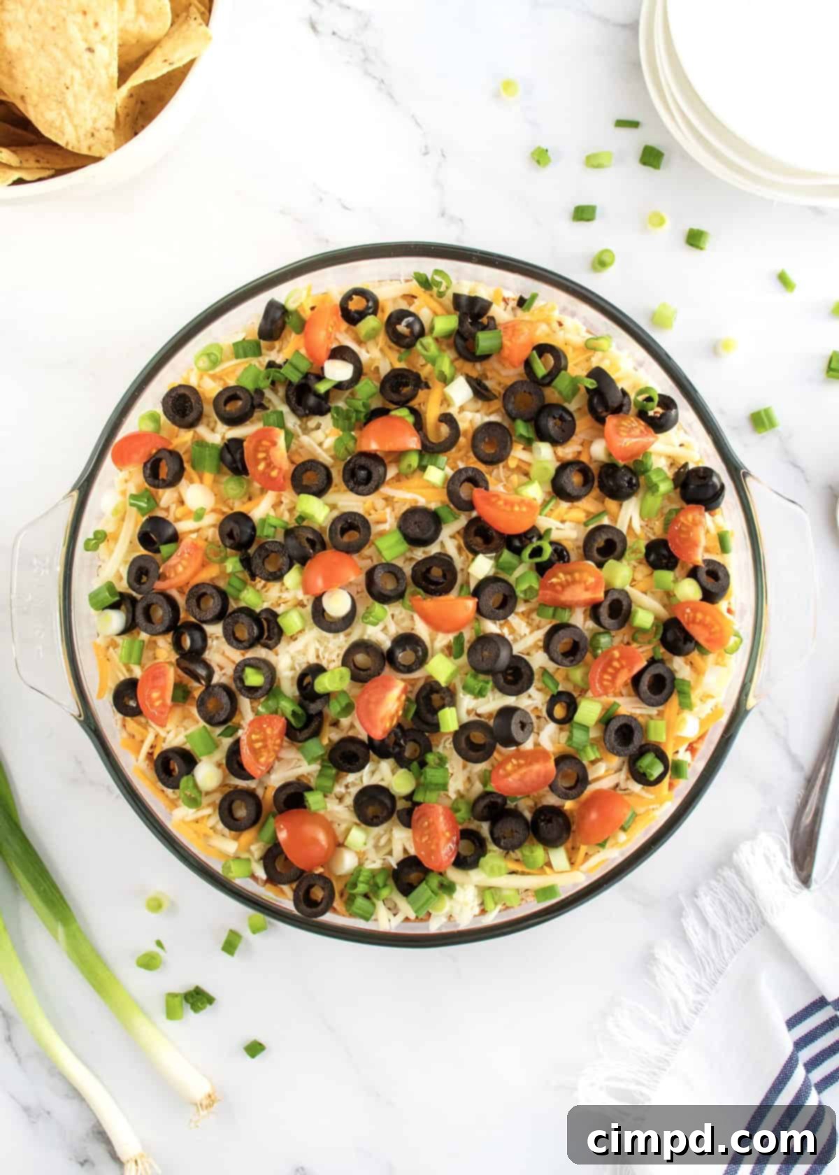 The finished 7-Layer Dip, garnished with cherry tomatoes and green onions, served in a glass dish with a bowl of tortilla chips on the side, ready for a party.
