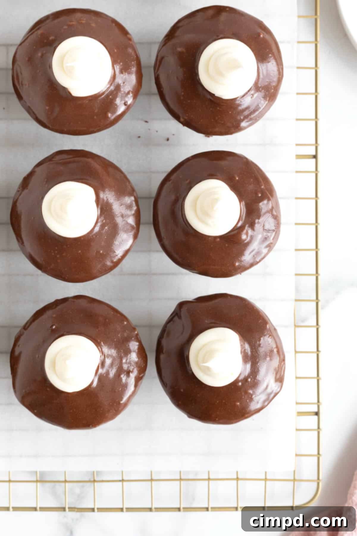 Six dark chocolate cupcakes with rich chocolate ganache and marshmallow cream topping, neatly arranged on a parchment-lined gold tone cooling rack.