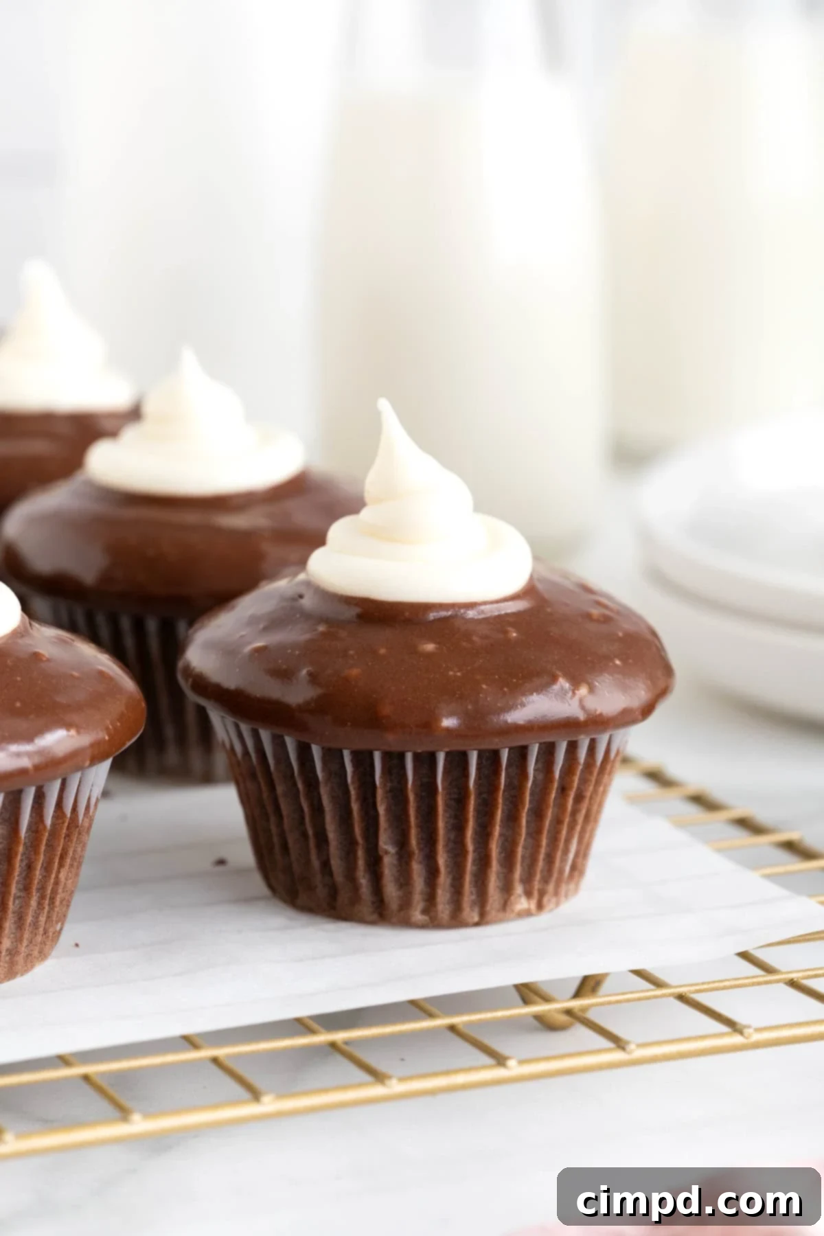 Five dark chocolate cupcakes, generously topped with chocolate ganache and a dollop of marshmallow cream, resting on a parchment-lined gold cooling rack.