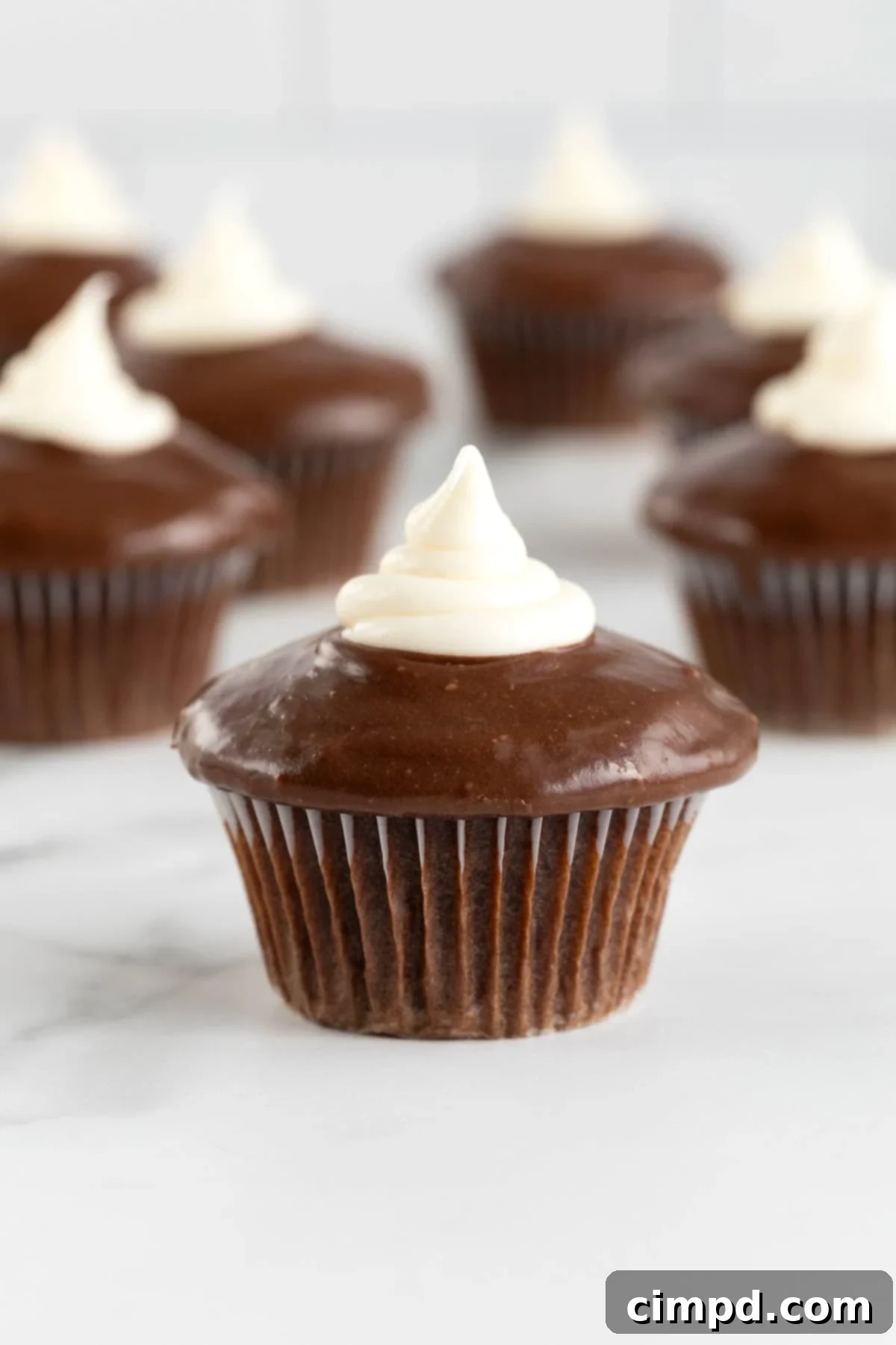 Five dark chocolate cupcakes topped with a swirl of chocolate ganache and a dollop of marshmallow cream, arranged on a white marble counter.