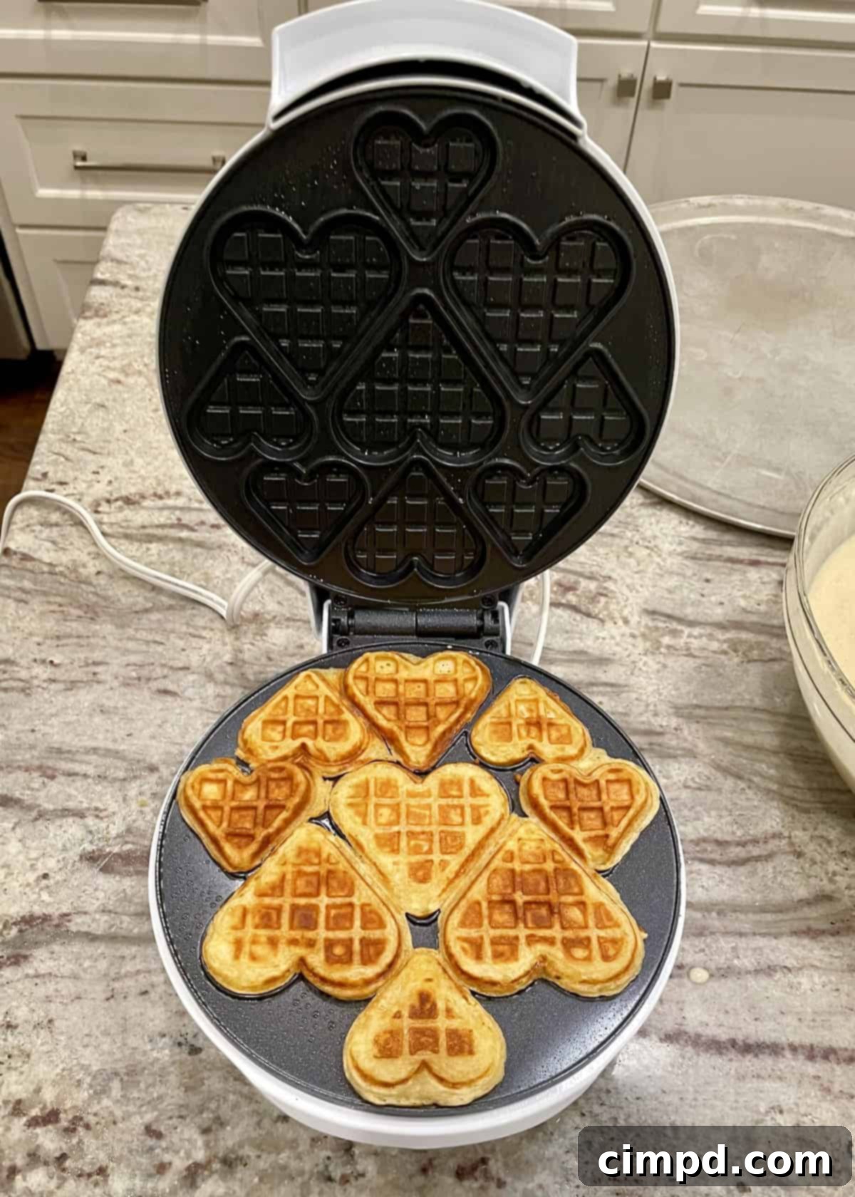 Heart-shaped waffles cooking in a mini waffle maker, part of the Valentine's Day Waffle Board preparation.