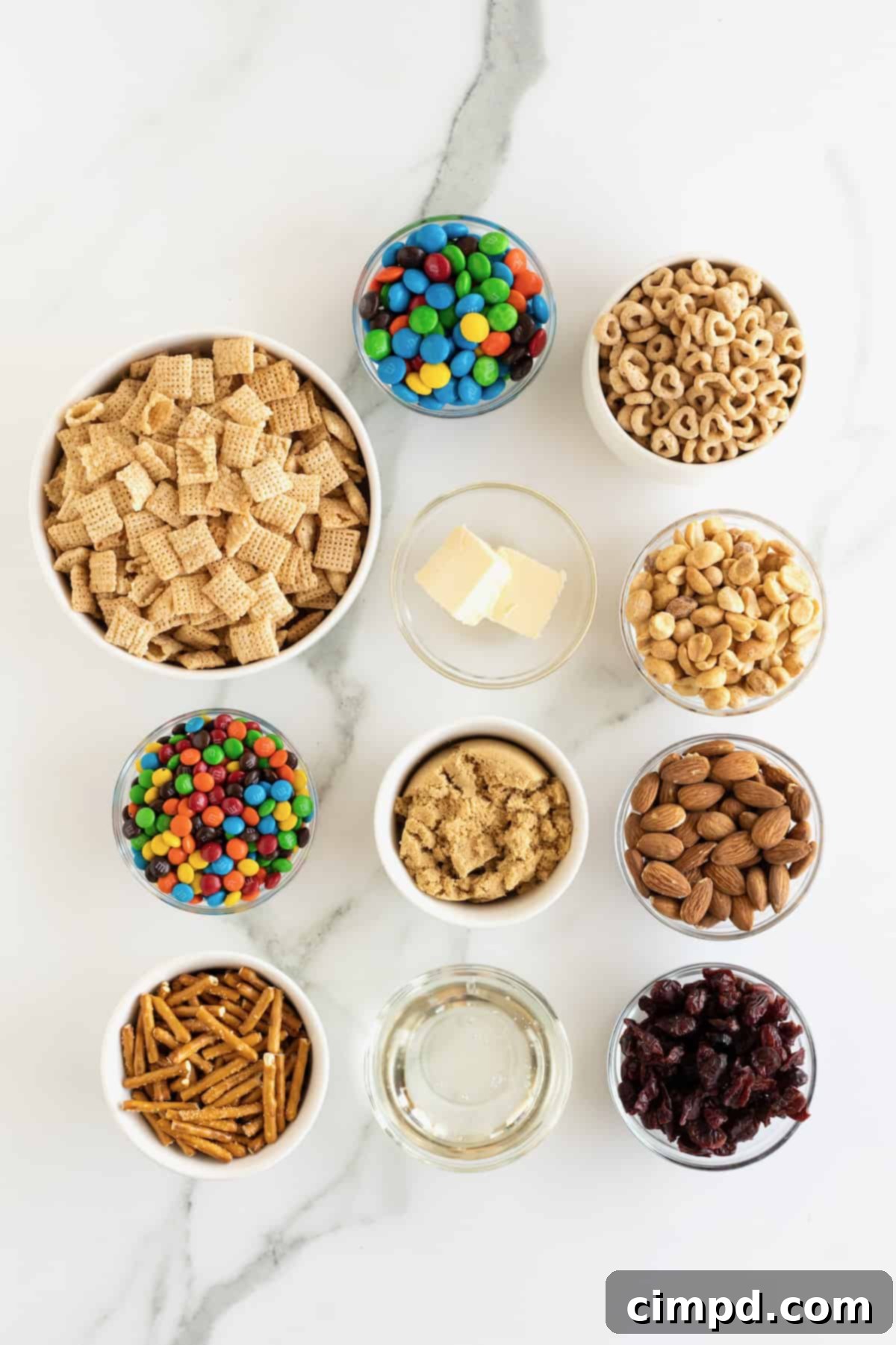 No-Bake Grab-and-Go Bars 5 Individual bowls filled with ingredients for no-bake snack bars: Chex cereal, Cheerios, M&Ms, M&M minis, roasted peanuts, almonds, and dried cranberries, alongside pretzel sticks.