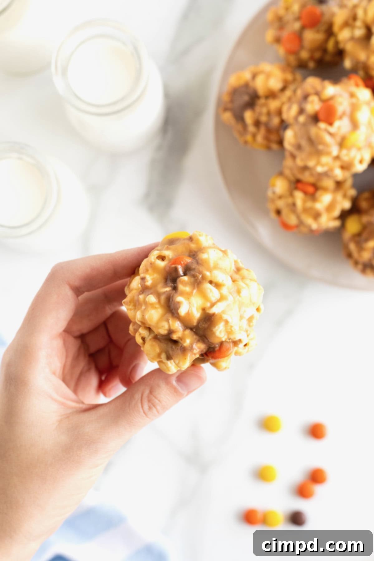 A hand holding a peanut butter popcorn ball in front of a large white serving plate of popcorn balls.
