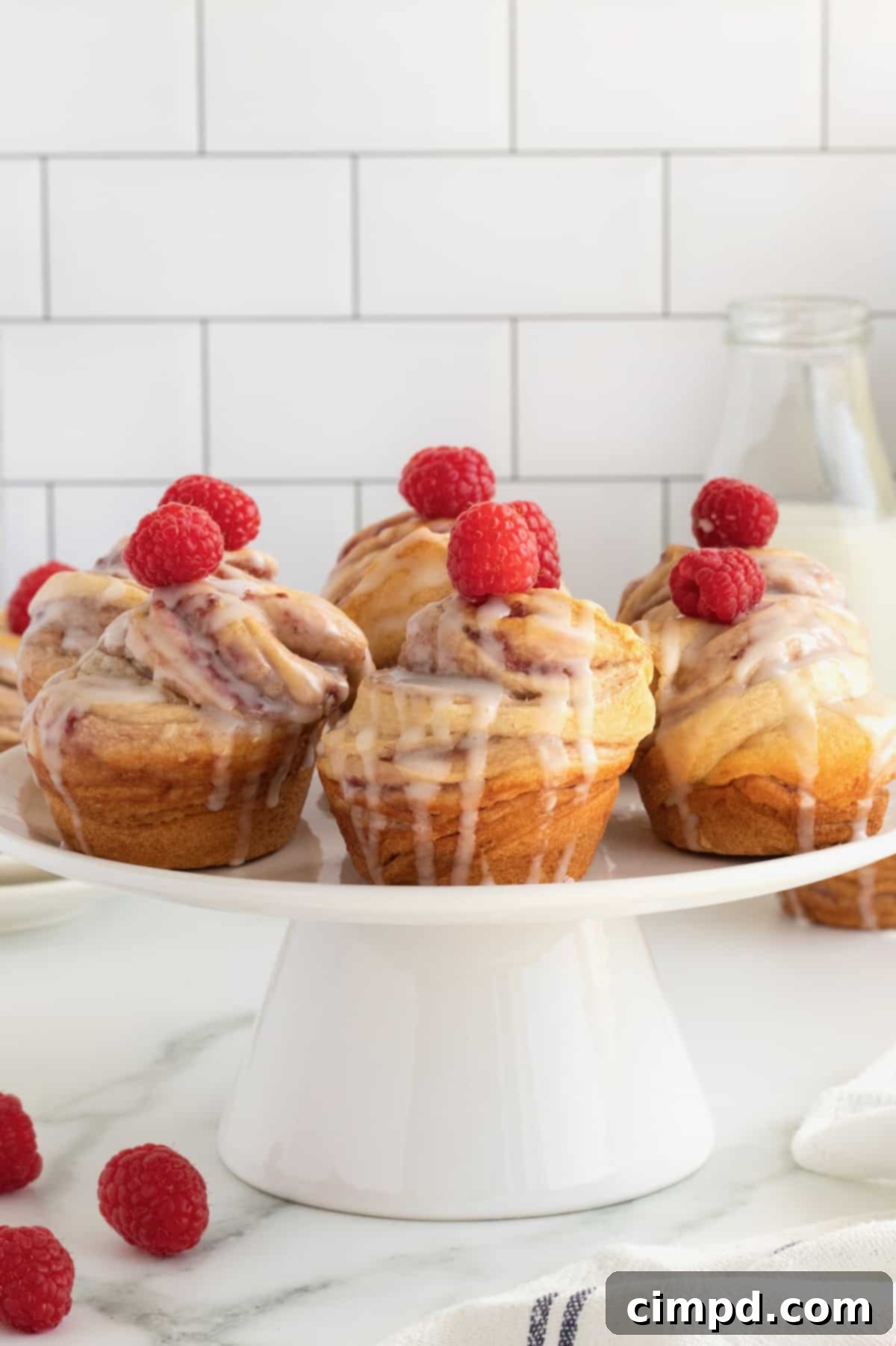 A beautiful cake stand filled with six glazed Raspberry Cream Cheese Cruffins, each crowned with a fresh raspberry, ready to be served.