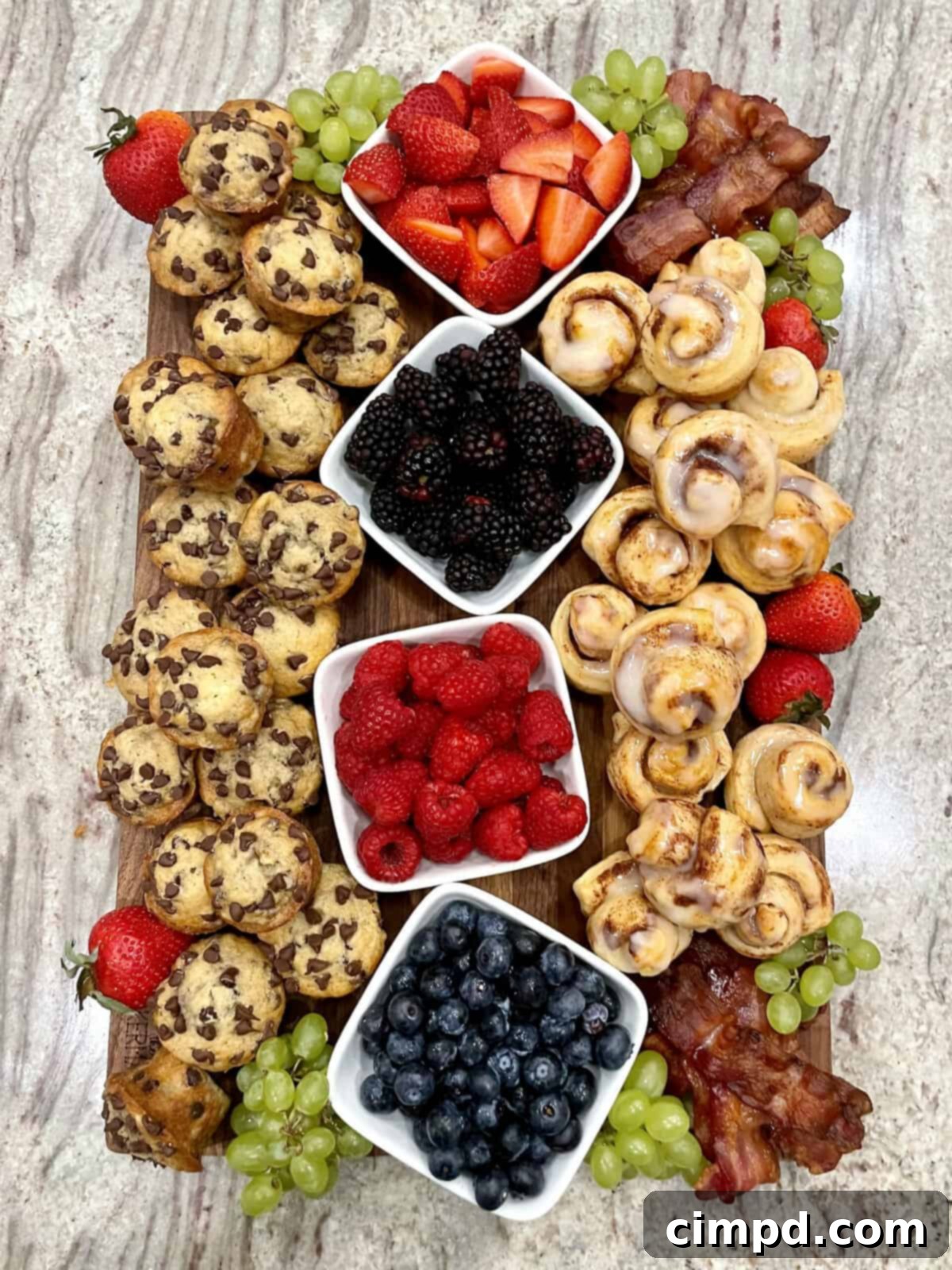 A vibrant Mini Muffin and Cinnamon Roll Breakfast Board by The BakerMama, featuring golden mini muffins, swirled cinnamon rolls, crispy bacon, and an assortment of fresh berries and grapes.