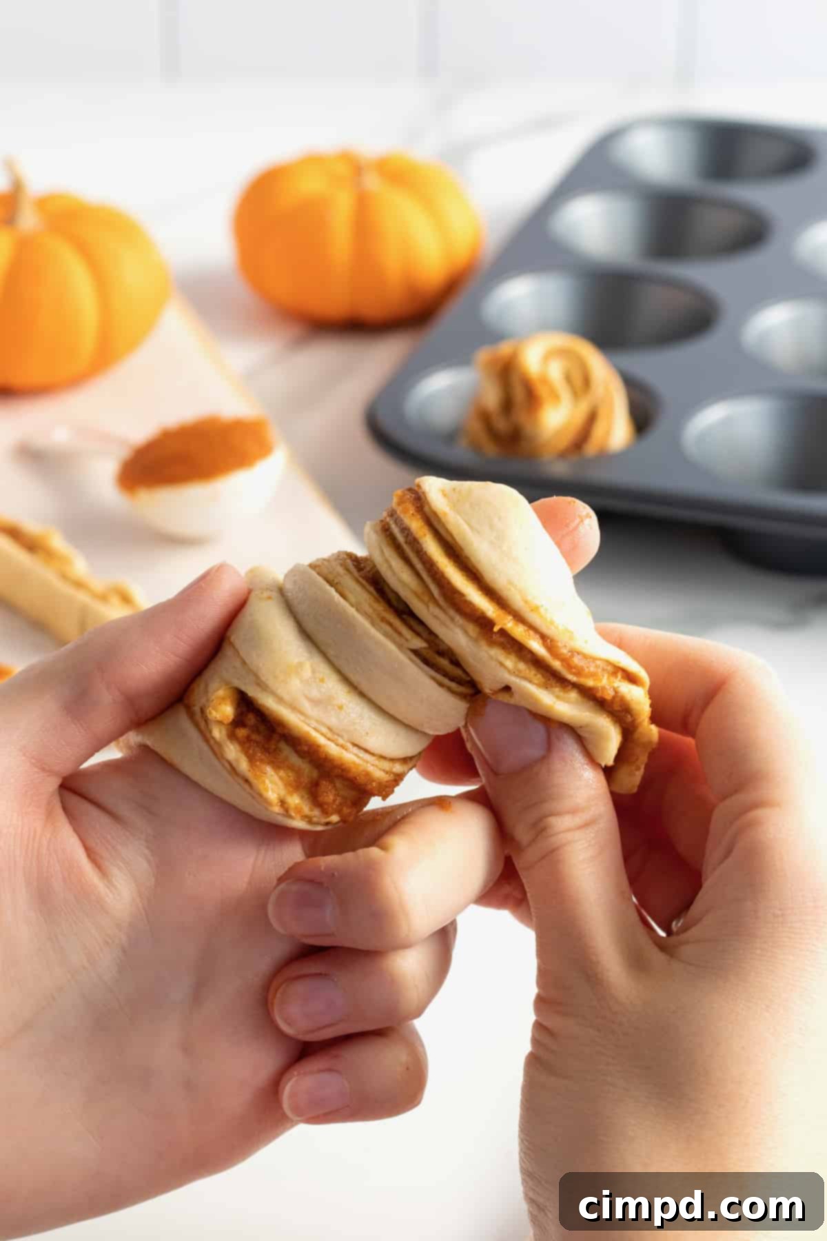 Spiced Pumpkin Cruffins 6 A hand demonstrating how to wrap a strip of pumpkin spice crescent roll dough around a finger to form the cruffin shape, showing the layered pastry.