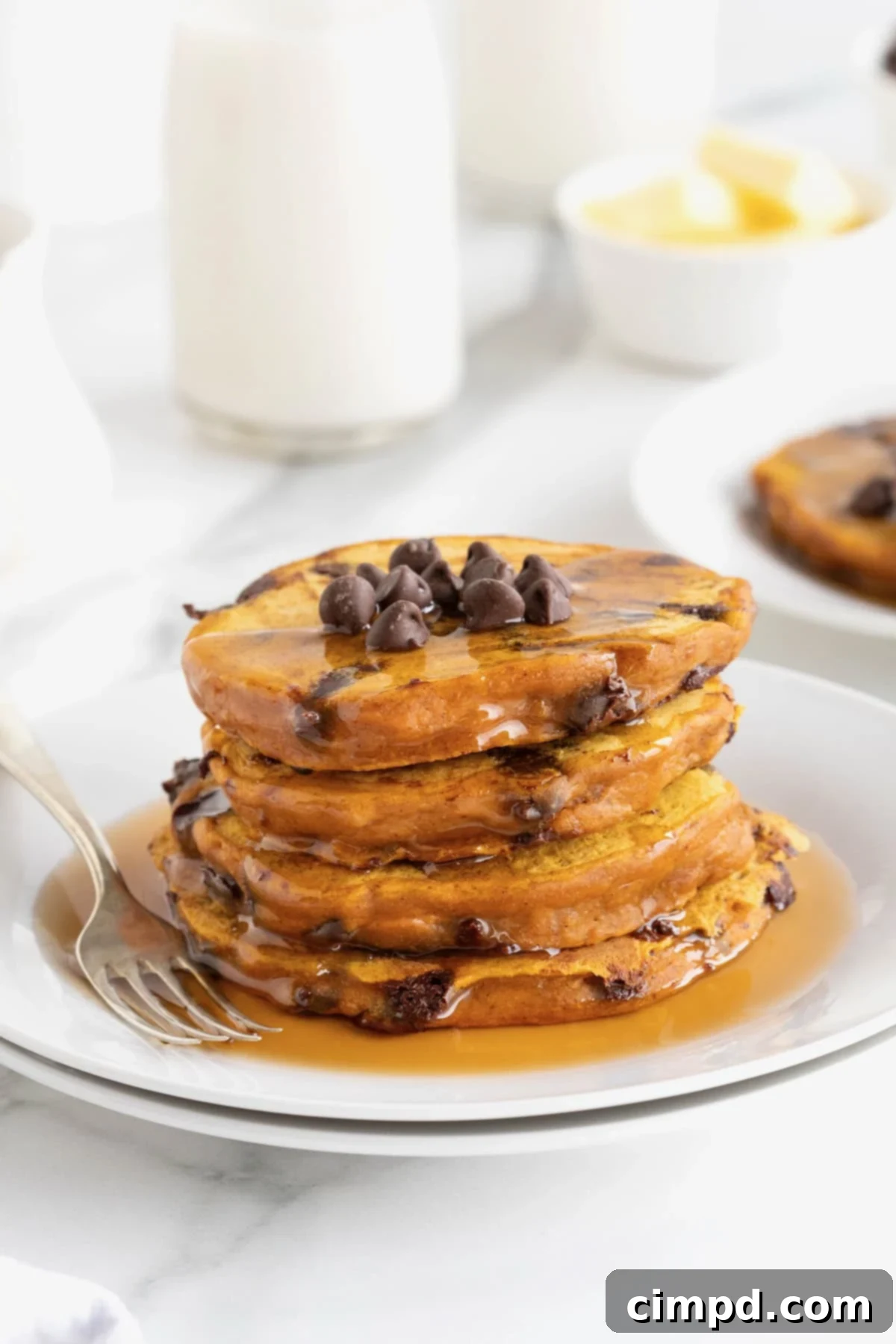 Spiced Pumpkin Chocolate Chip Pancakes 2 Four fluffy pumpkin chocolate chip pancakes stacked high, generously drizzled with syrup on a pristine white plate, ready to be enjoyed.