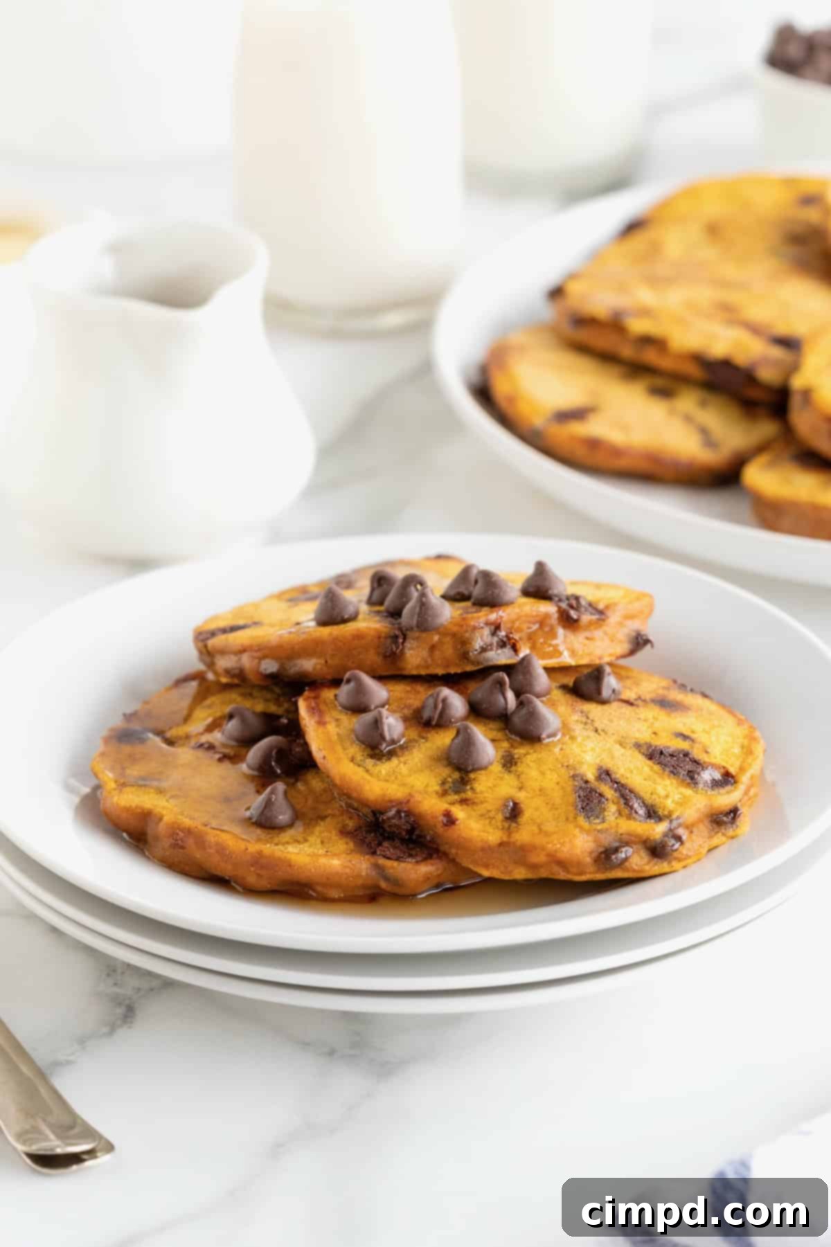 Spiced Pumpkin Chocolate Chip Pancakes 11 A close-up of three golden pumpkin pancakes, beautifully topped with melted chocolate chips, resting on a stack of pristine white plates, with more pancakes blurred in the background.