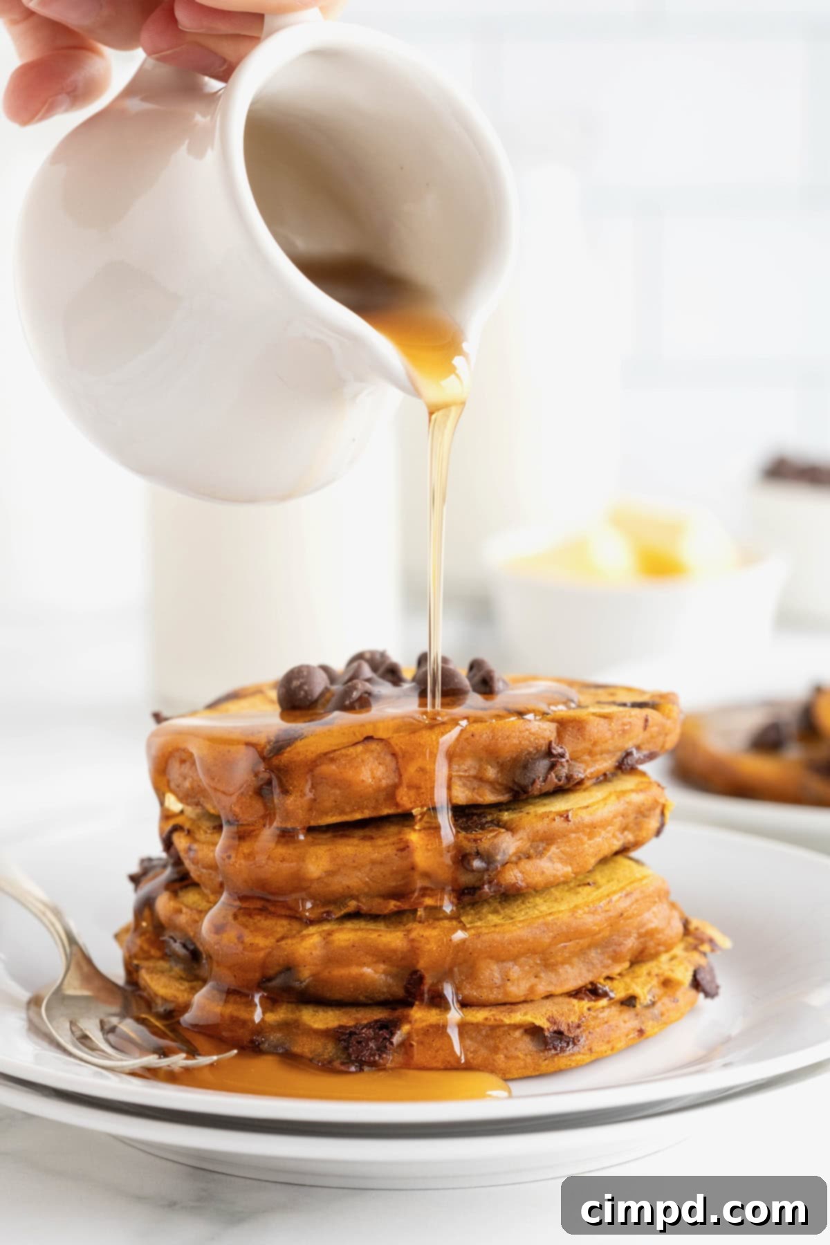 Spiced Pumpkin Chocolate Chip Pancakes 4 Golden maple syrup being generously poured from a small white carafe over a tower of four pumpkin pancakes, adorned with glistening chocolate chips, on a white plate.