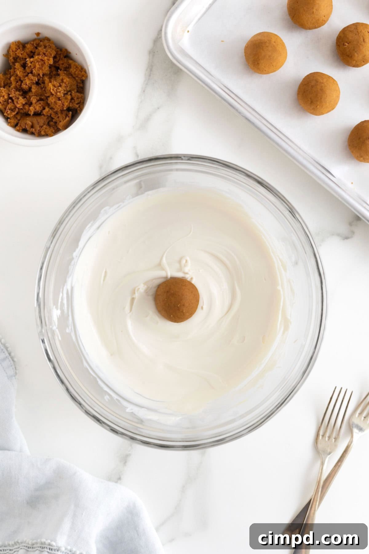 A pumpkin cake ball being carefully coated in a glass bowl of melted white candy coating, ensuring an even layer.