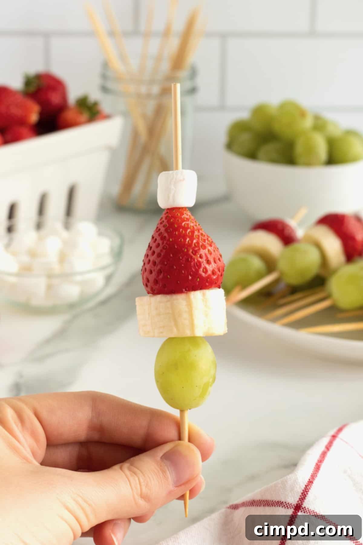 Close-up of a Grinch Fruit Skewer, showcasing the red strawberry hat, white marshmallow pom-pom, banana face, and green grape body.