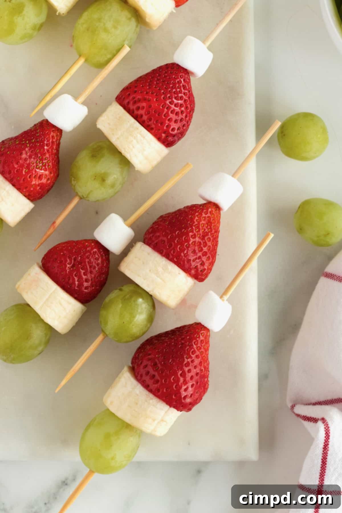 Children's hands assembling Grinch Fruit Skewers, showing the step-by-step process of threading fruit onto bamboo sticks.