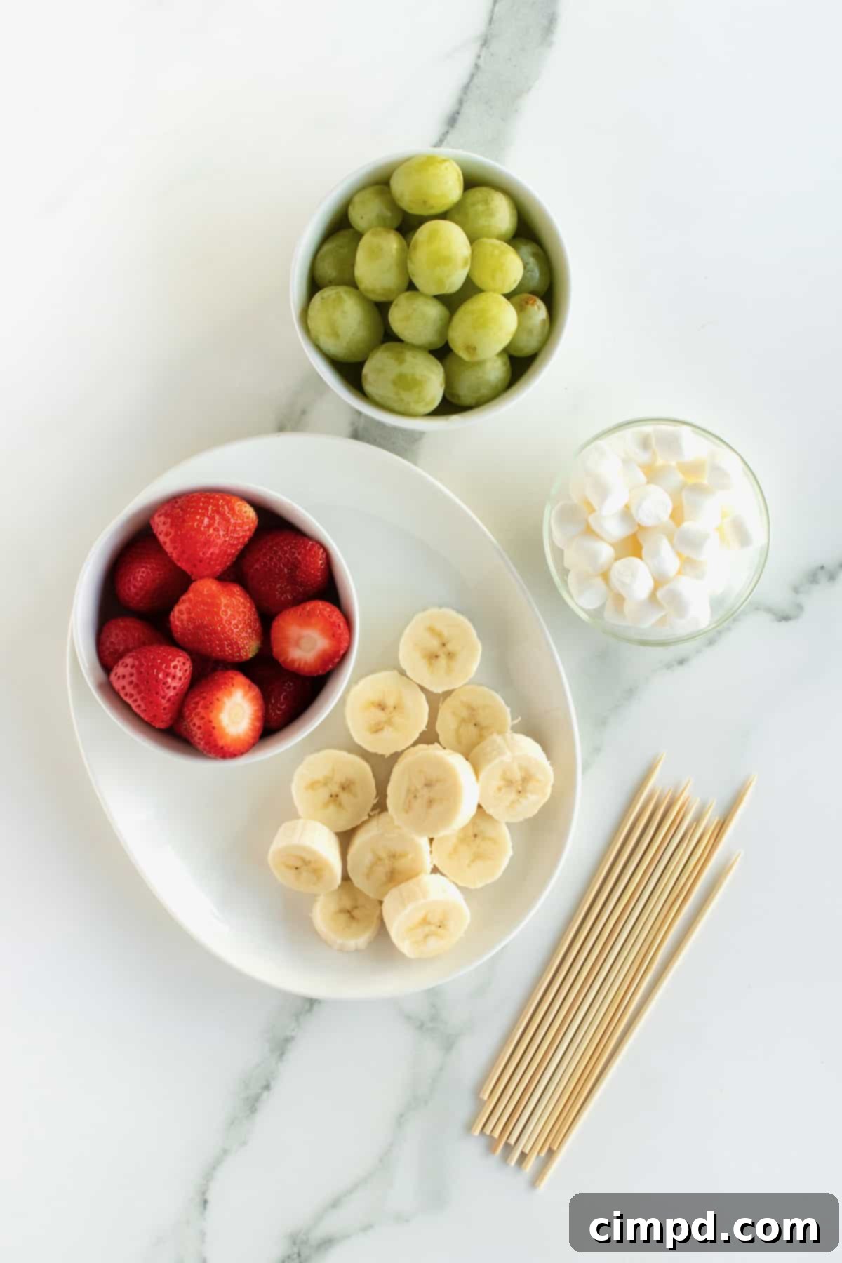 Ingredients for Grinch Fruit Skewers laid out: mini marshmallows, strawberries, banana slices, green grapes, and bamboo skewers.