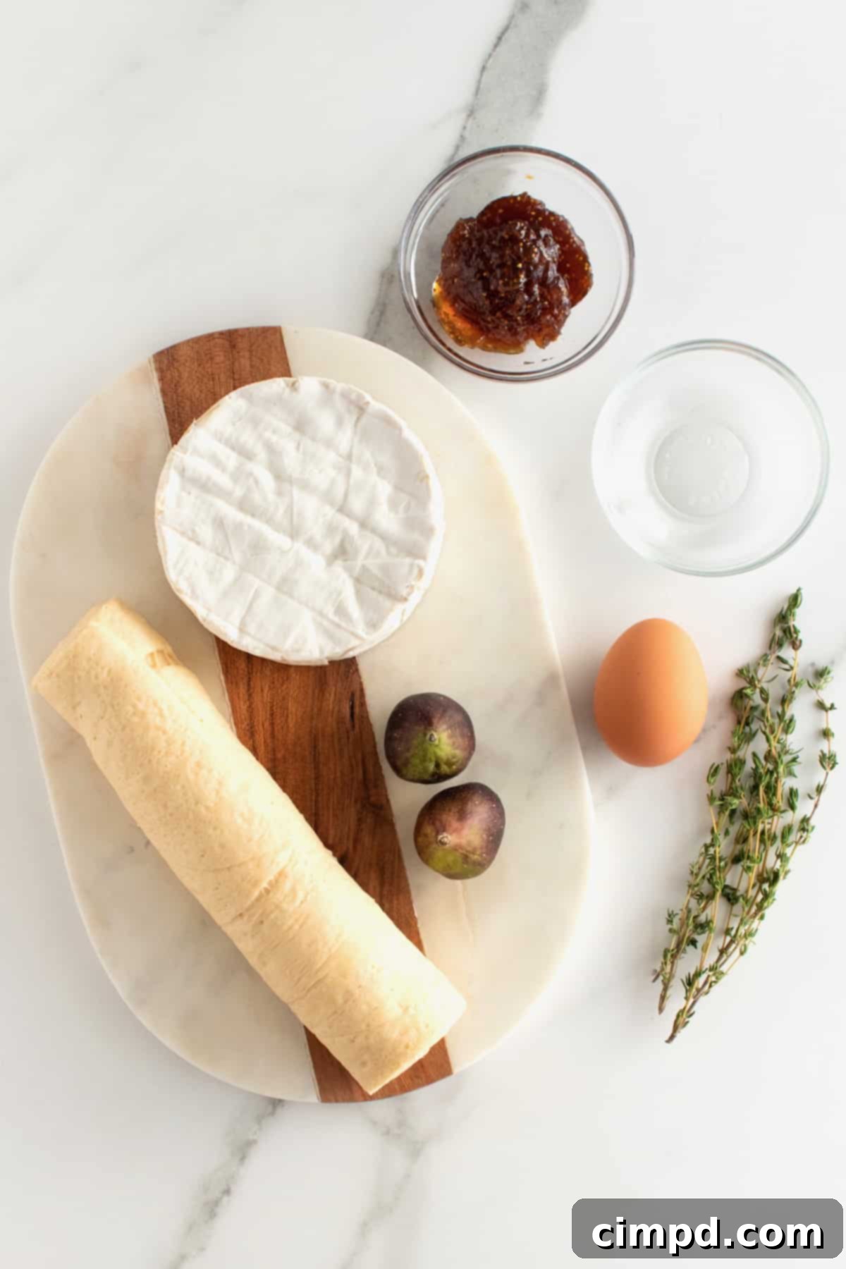 Ingredients for baked brie with fig jam laid out on a kitchen counter: crescent dough, brie, fig jam, fresh figs, egg, and thyme.