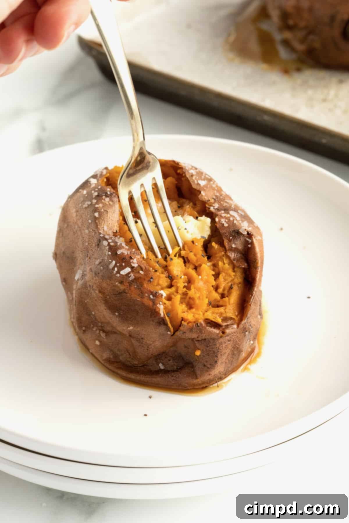 Perfect Baked Sweet Potatoes 6 A sweet potato on a white plate. The potato is split open and topped with butter, salt and pepper. There is a metal fork sticking out of the potato.