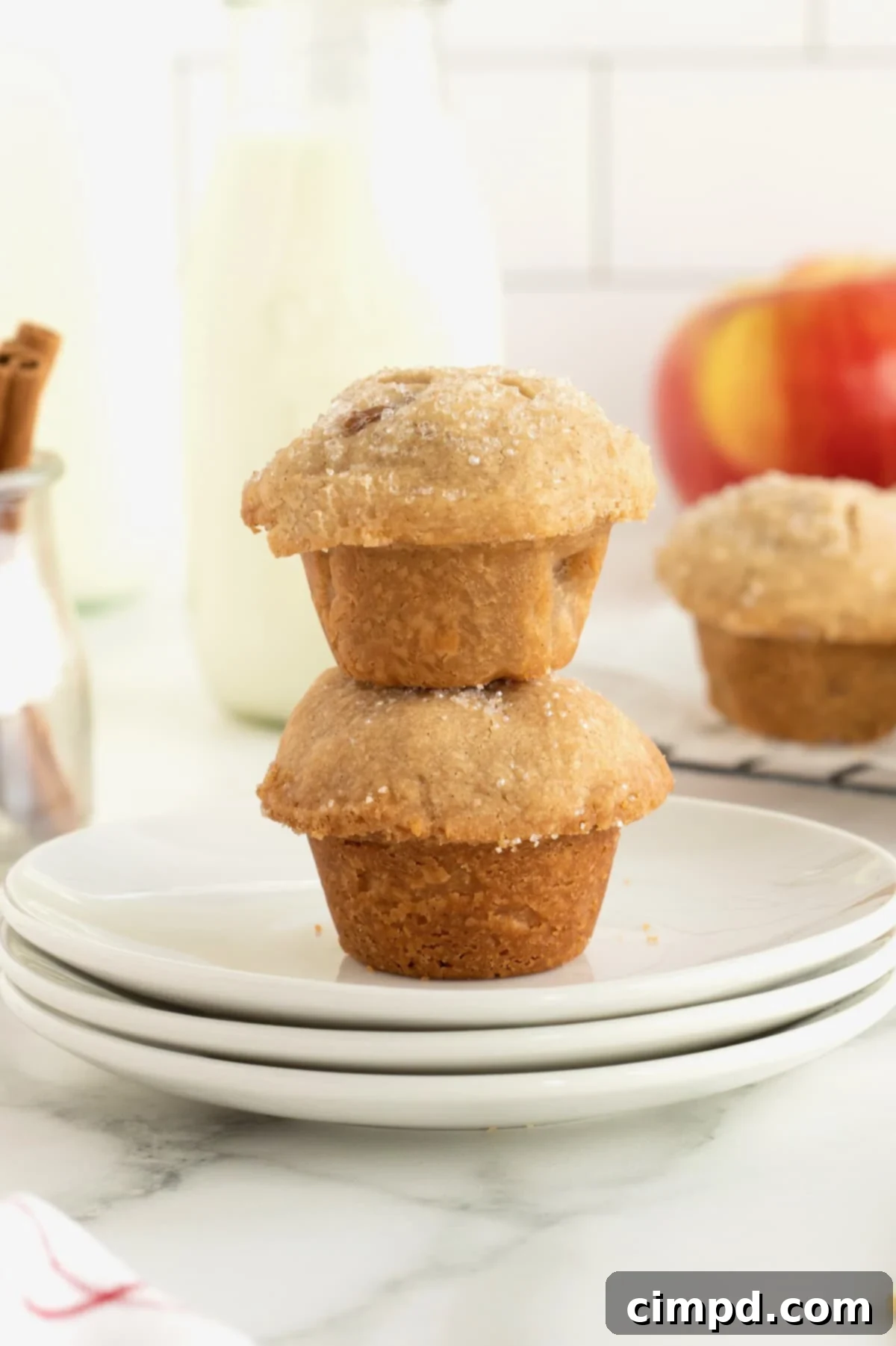 Two bite-sized apple pie bites stacked on a small white plate with apples and a glass of milk in the background.