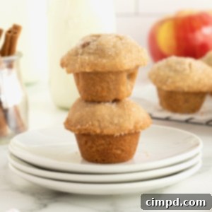Two bite-sized apple pie bites stacked on a small white plate with apples and a glass of milk in the background.