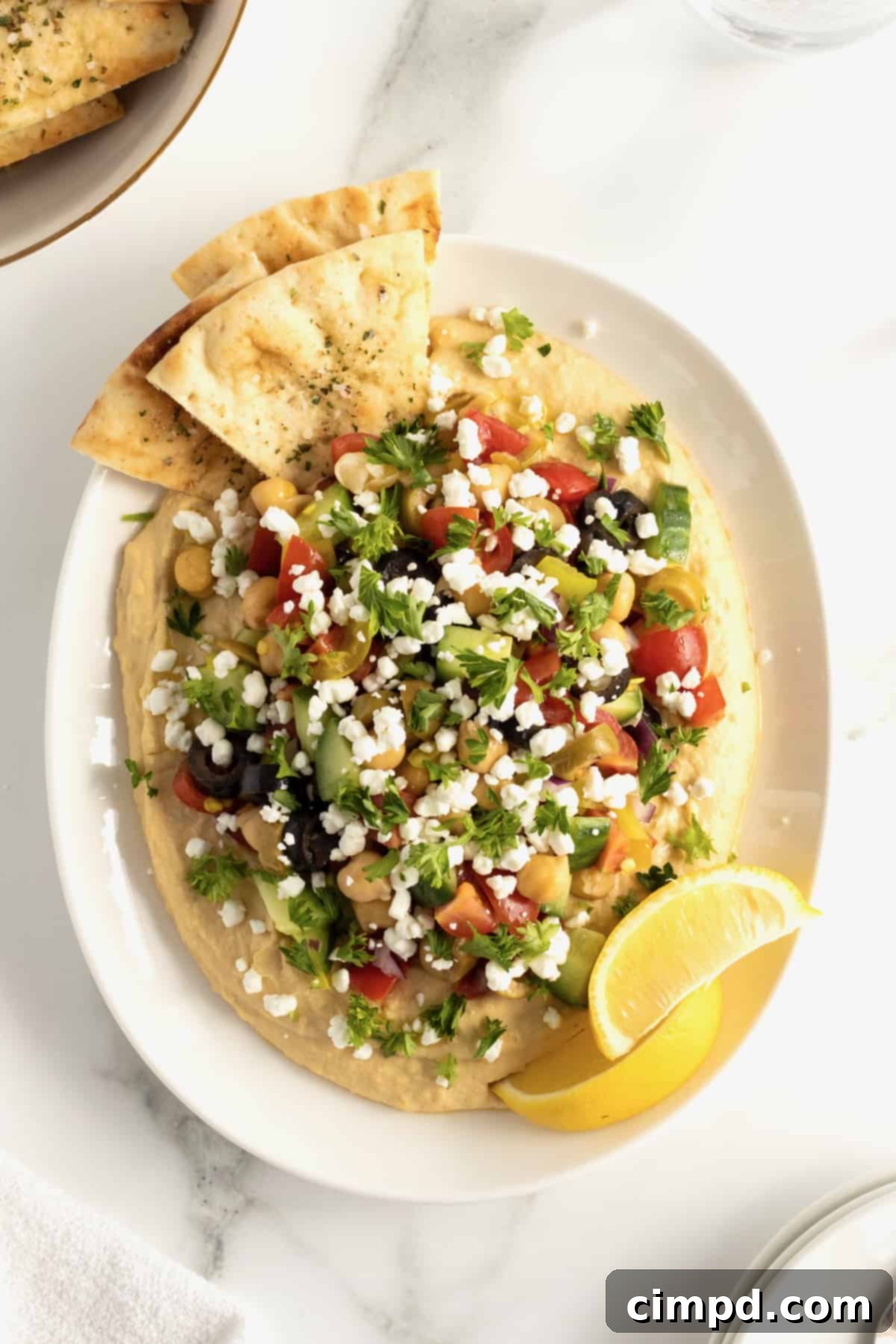 A vibrant white ceramic serving dish filled with creamy hummus, generously topped with a colorful medley of chopped tomatoes, olives, cucumber, chickpeas, red onion, and bright pepperoncini peppers. Triangles of baked pita bread are artfully arranged around the dish, inviting a delicious dip.