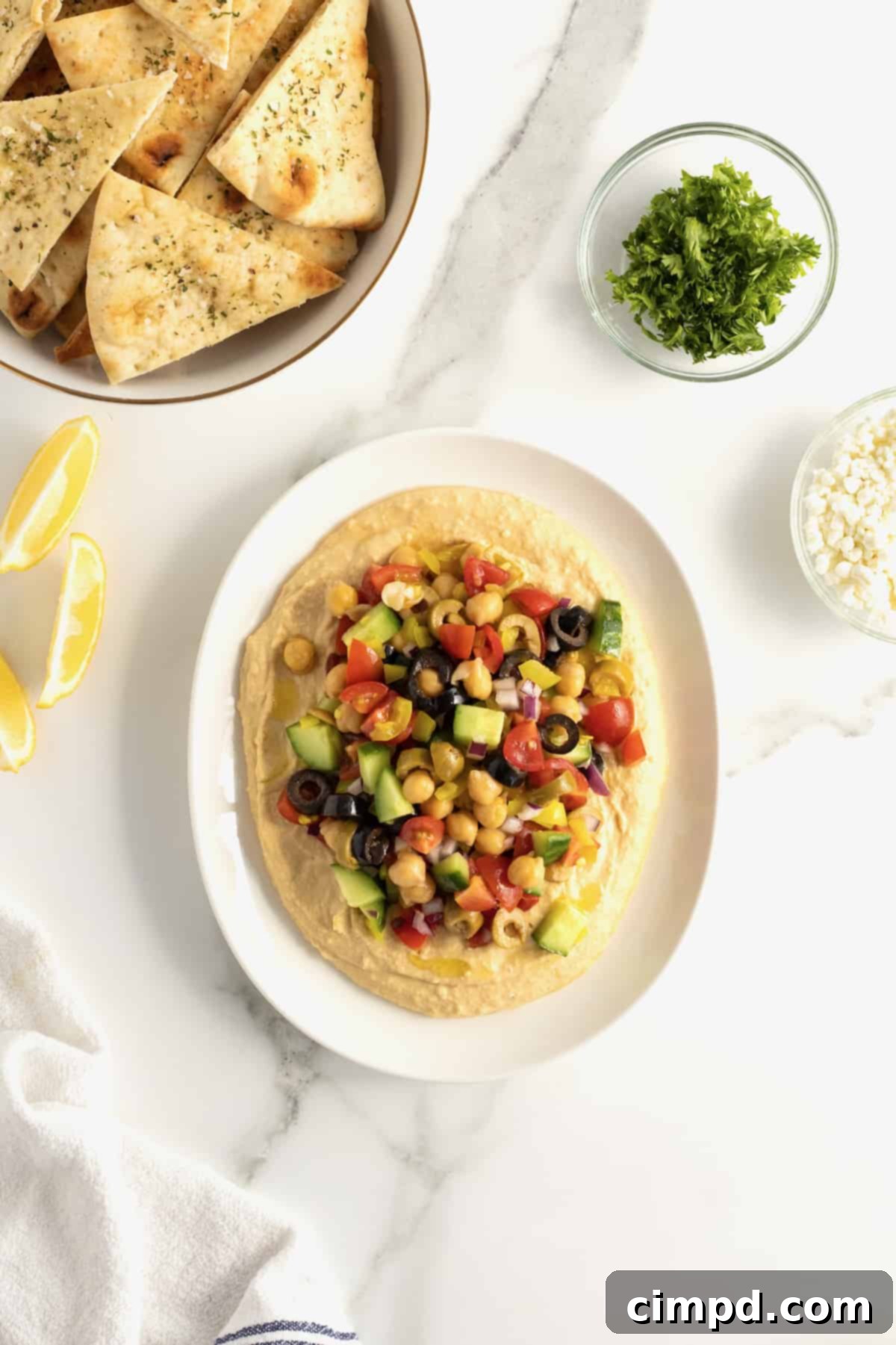 A white ceramic serving dish featuring creamy hummus generously topped with a vibrant mix of chopped tomatoes, olives, cucumber, chickpeas, red onion, and pepperoncini peppers. A separate white bowl filled with baked pita chips sits invitingly beside the hummus platter.