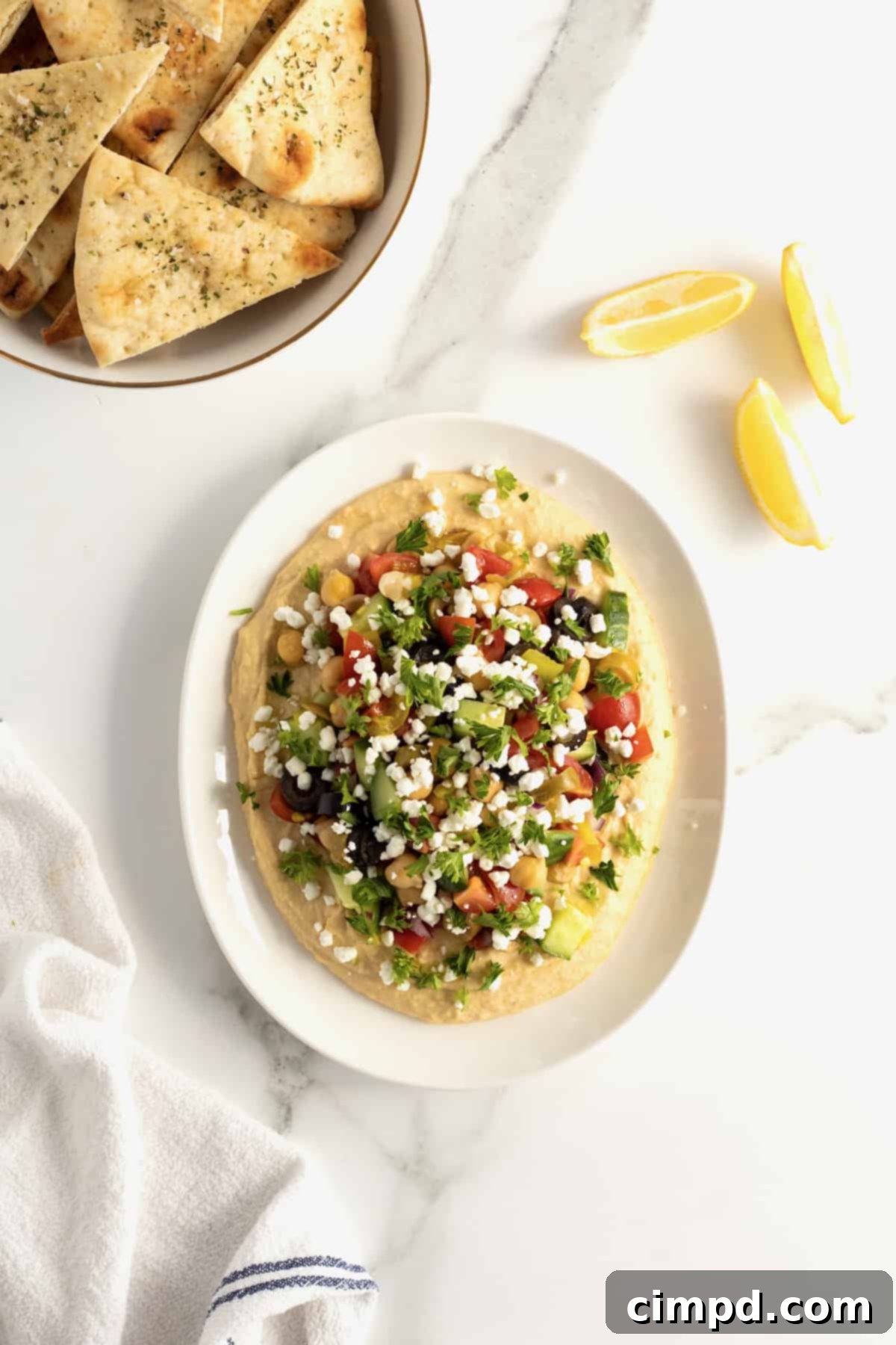 A beautifully presented white ceramic serving dish of loaded hummus, vibrant with chopped tomatoes, olives, cucumber, chickpeas, red onion, pepperoncini peppers, crumbled feta, and fresh parsley. It is accompanied by bright lemon wedges and a pile of golden baked pita chips, ready to be enjoyed.