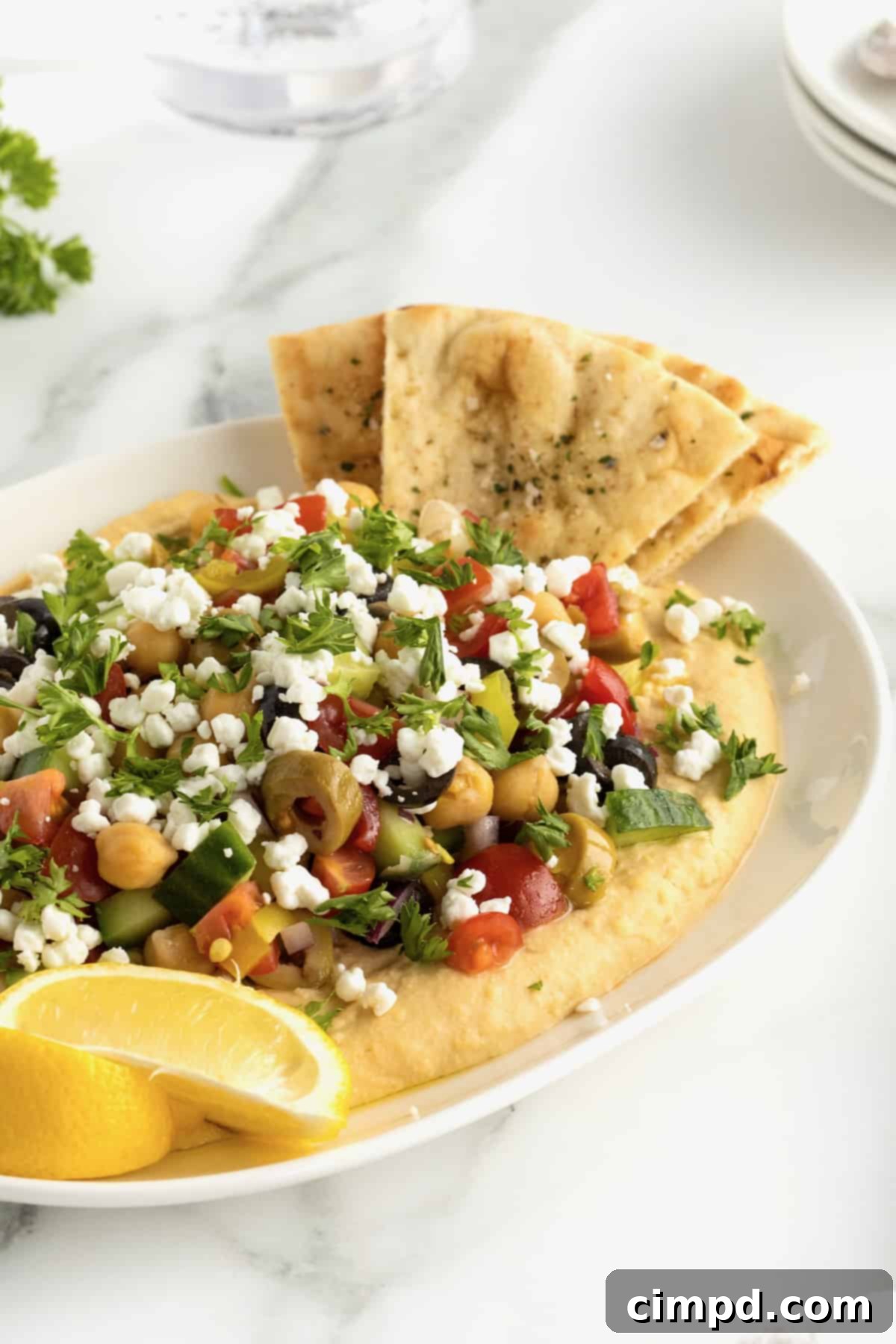 A beautifully presented serving dish of Loaded Hummus by The BakerMama, featuring creamy hummus generously topped with colorful fresh vegetables, feta, and parsley, ready to be enjoyed.