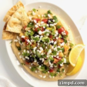 Hummus topped with tomatoes, olives, cucumber, chickpeas, red onion and pepperoncini peppers in a white ceramic serving dish with pita triangles.