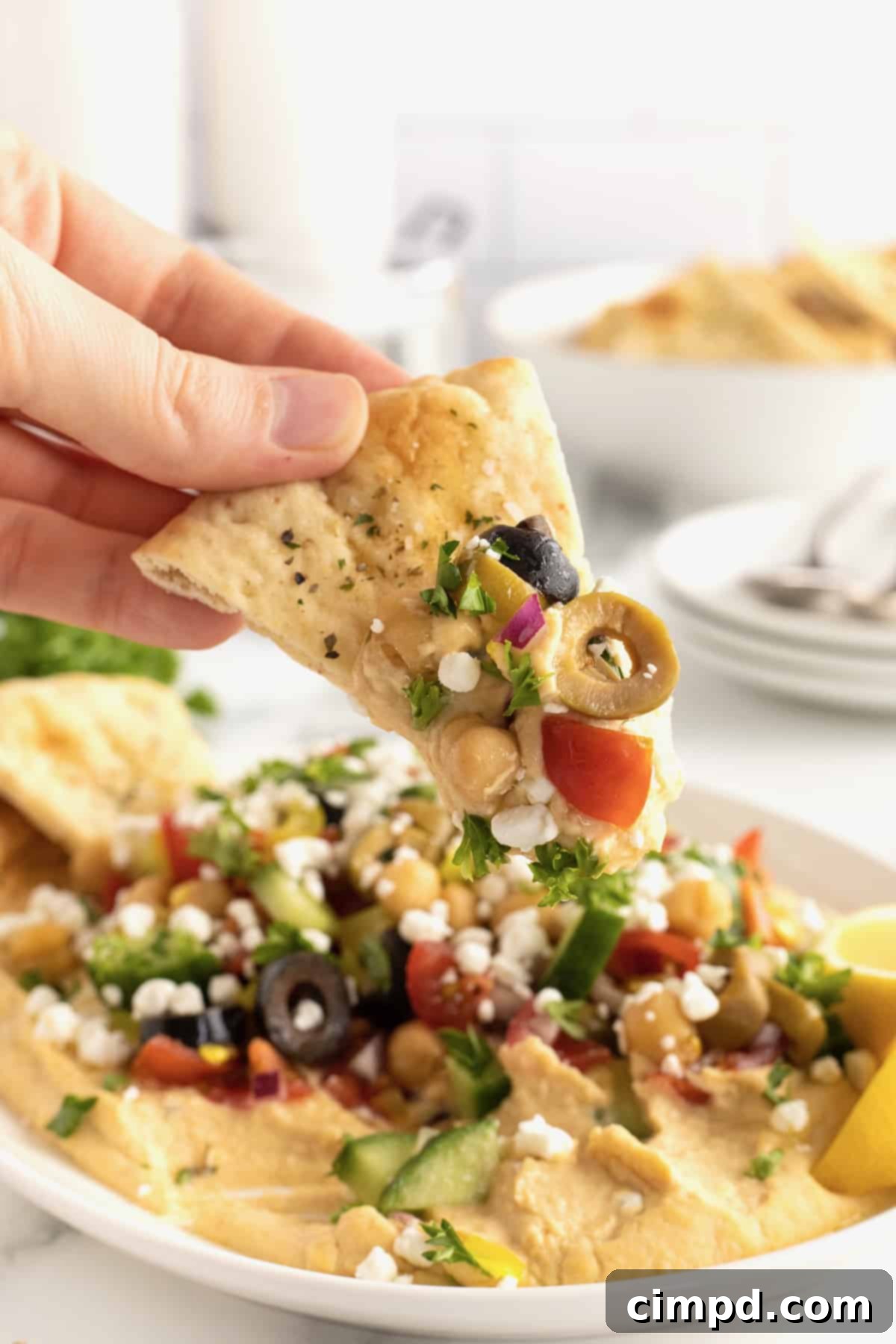 A close-up shot of a golden-brown pita chip, perfectly loaded with a generous scoop of hummus and large, visible chunks of ripe tomato, dark olive, and creamy feta cheese, highlighting the rich texture and fresh ingredients.