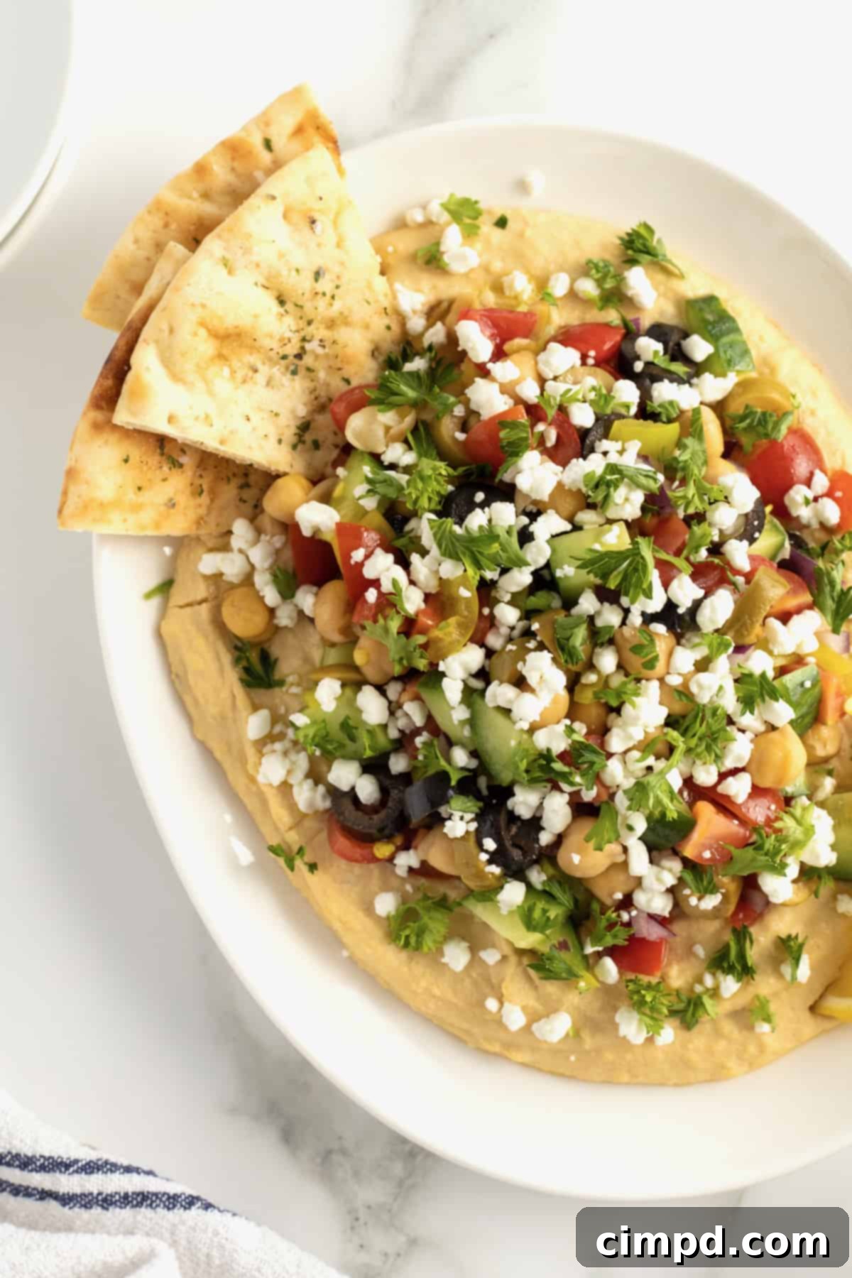A beautifully presented white ceramic serving dish, brimming with creamy hummus and lavishly adorned with a colorful array of chopped tomatoes, briny olives, crisp cucumber, tender chickpeas, finely diced red onion, and vibrant pepperoncini peppers. Fluffy pita triangles are arranged alongside, ready for dipping.