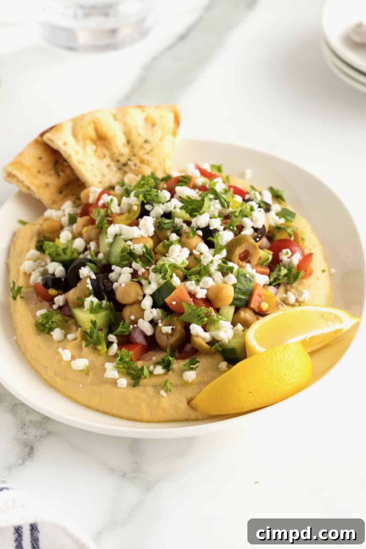 A white ceramic serving dish holds a generous portion of creamy hummus, artfully topped with a colorful mix of chopped tomatoes, briny olives, crisp cucumber, tender chickpeas, finely diced red onion, and zesty pepperoncini peppers. Lemon wedges and baked pita triangles are arranged invitingly alongside the dish.