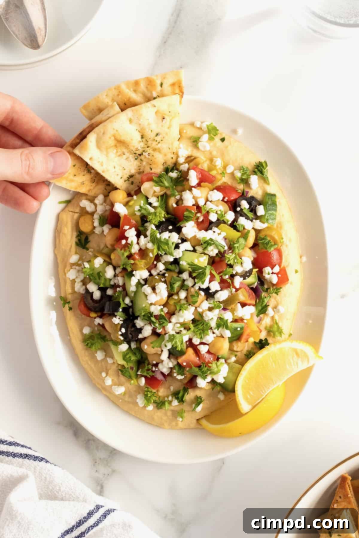 A hand reaches for a pita triangle, poised to scoop up a generous portion of the loaded hummus. The white ceramic serving dish is piled high with creamy hummus and a vibrant array of toppings including chopped tomatoes, briny olives, crisp cucumber, tender chickpeas, finely diced red onion, and zesty pepperoncini peppers.