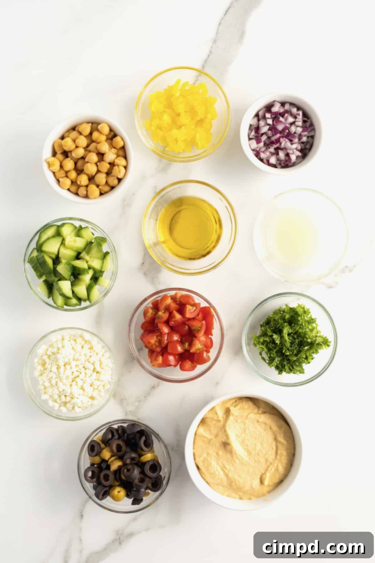 An array of fresh ingredients for loaded hummus, including vibrant chopped tomatoes, briny olives, crisp cucumber, tender chickpeas, finely diced red onion, and zesty pepperoncini peppers, neatly arranged in glass and ceramic dishes, alongside a bowl of plain hummus in a white ceramic dish.