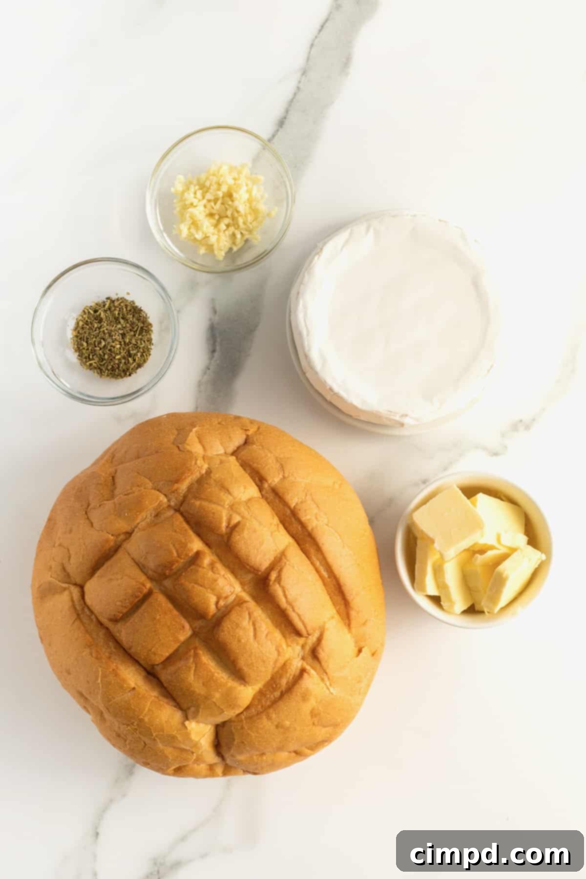 A bread bowl on a marble countertop, next to a wheel of brie and glass bowls filled with garlic, butter, and Italian seasoning.