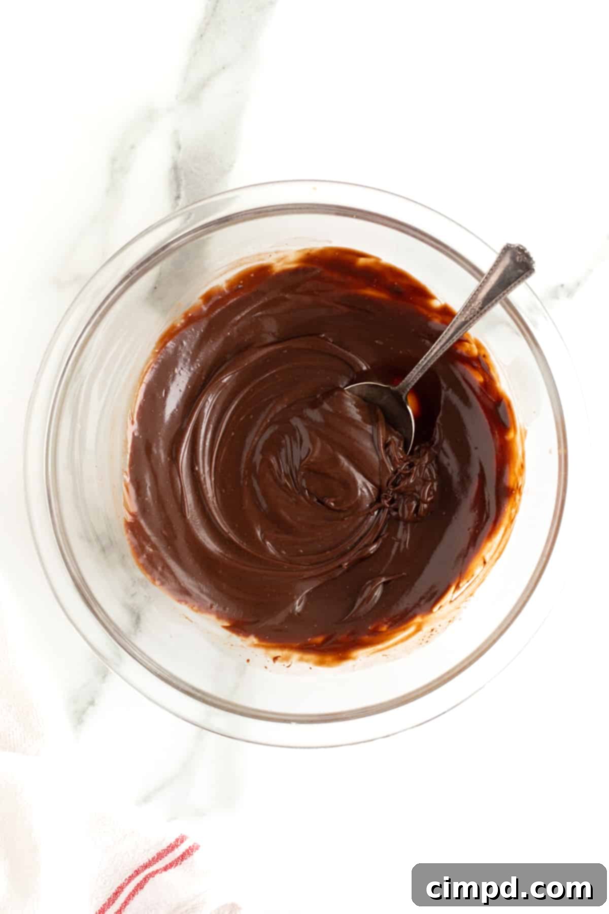 A pristine clear glass bowl holds a generous portion of freshly made chocolate ganache, with a gleaming metal spoon resting inside, ready to be sampled. The ganache boasts a perfect, smooth, and reflective surface.