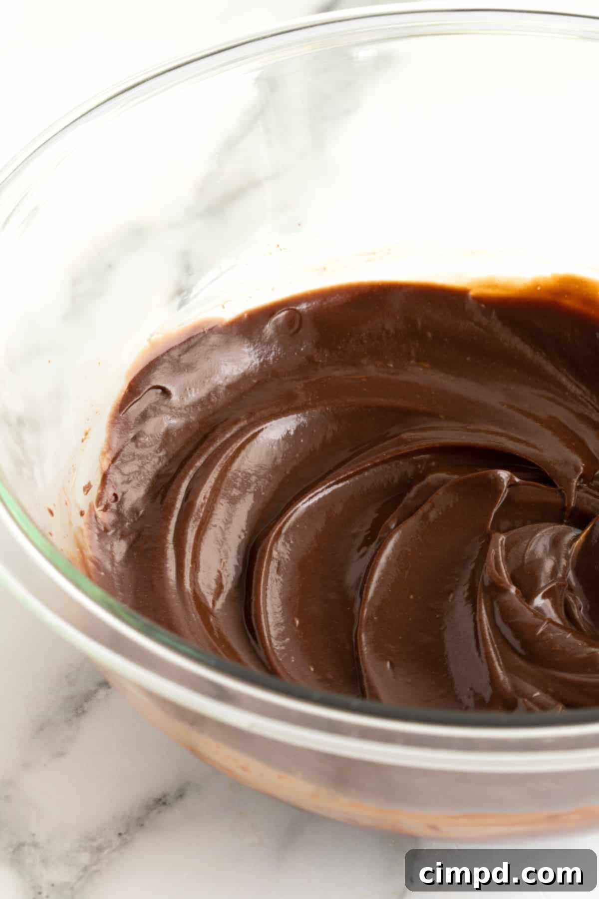 A clear glass bowl filled with glossy chocolate ganache, sealed tightly with plastic wrap pressed directly onto its surface, ready for refrigeration to maintain freshness.