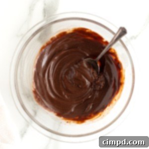 Close-up of smooth chocolate ganache in a clear glass bowl, with a spoon resting in it, ready for use as a delectable topping or filling.