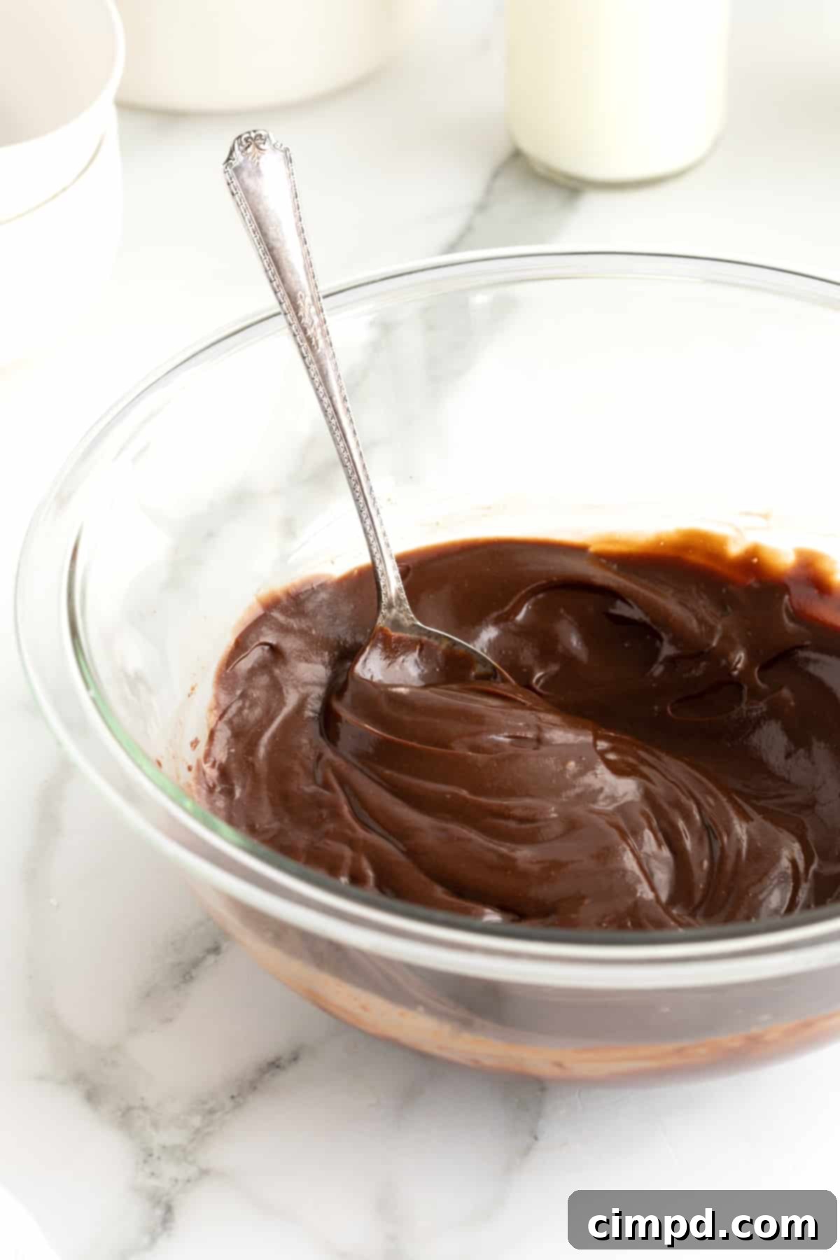 A glistening chocolate ganache fills a clear glass bowl, reflecting the light and promising rich flavor. A metal spoon is gently placed inside, inviting a taste of its smooth, creamy texture.