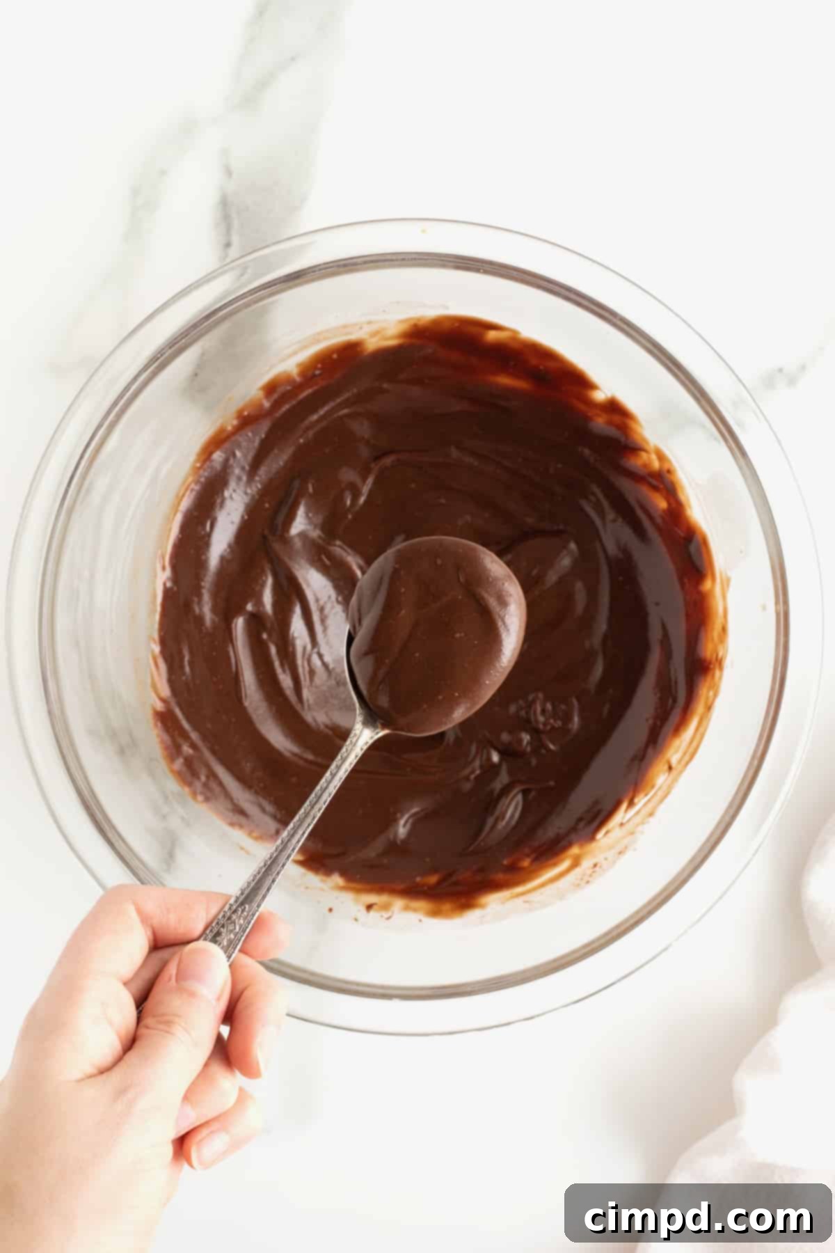 A close-up view of a clear glass bowl filled with rich, dark chocolate ganache. A hand holds a spoonful of the luscious ganache just above the bowl, showcasing its smooth, pourable consistency.