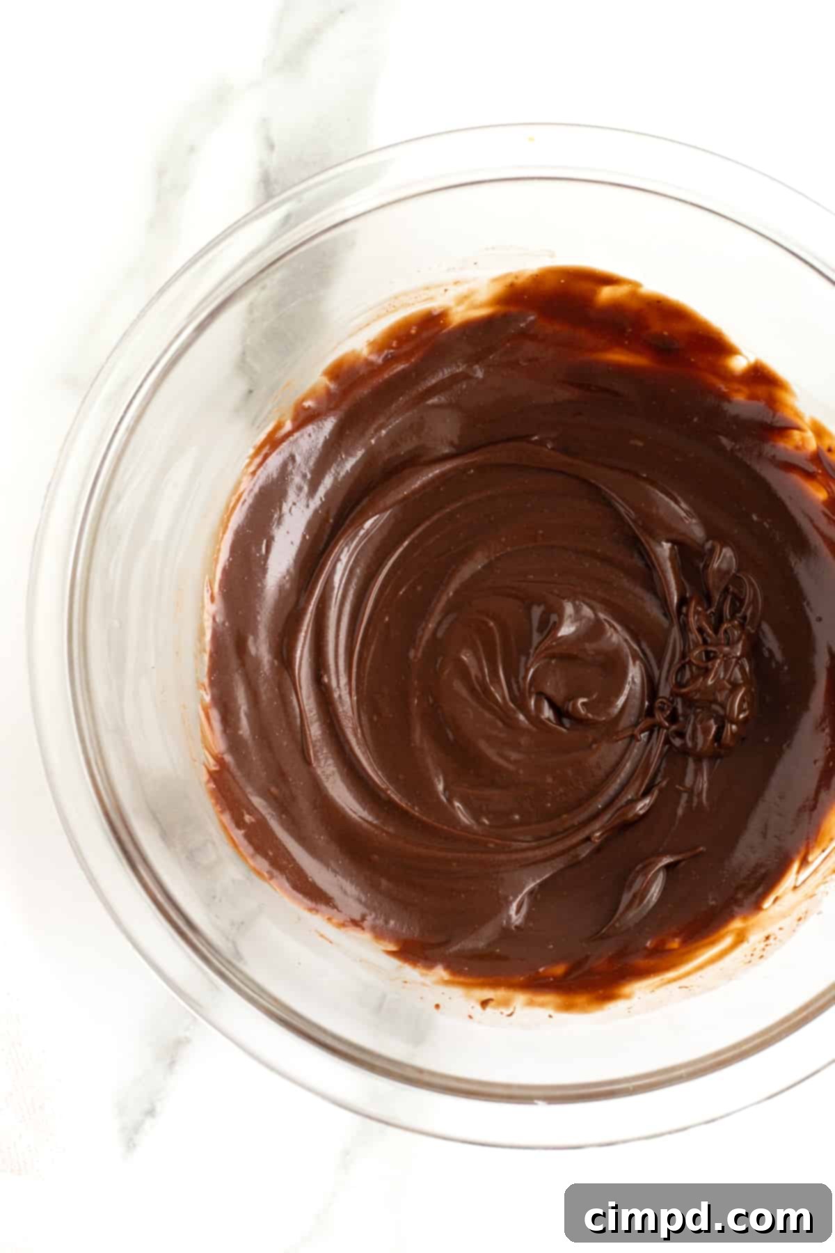 A clear glass bowl filled with smooth, dark chocolate ganache, resting on a clean white surface. The ganache has a beautiful, glossy sheen, indicating its perfect, velvety texture.