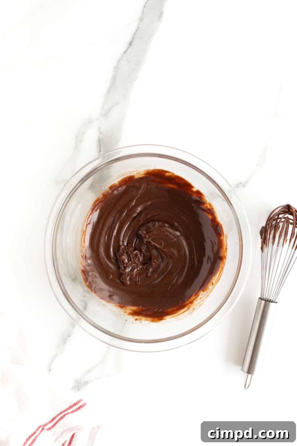 A clear glass bowl filled with freshly prepared chocolate ganache, smooth and glossy. A metal whisk rests beside the bowl, hinting at the effort to achieve such a perfect consistency.