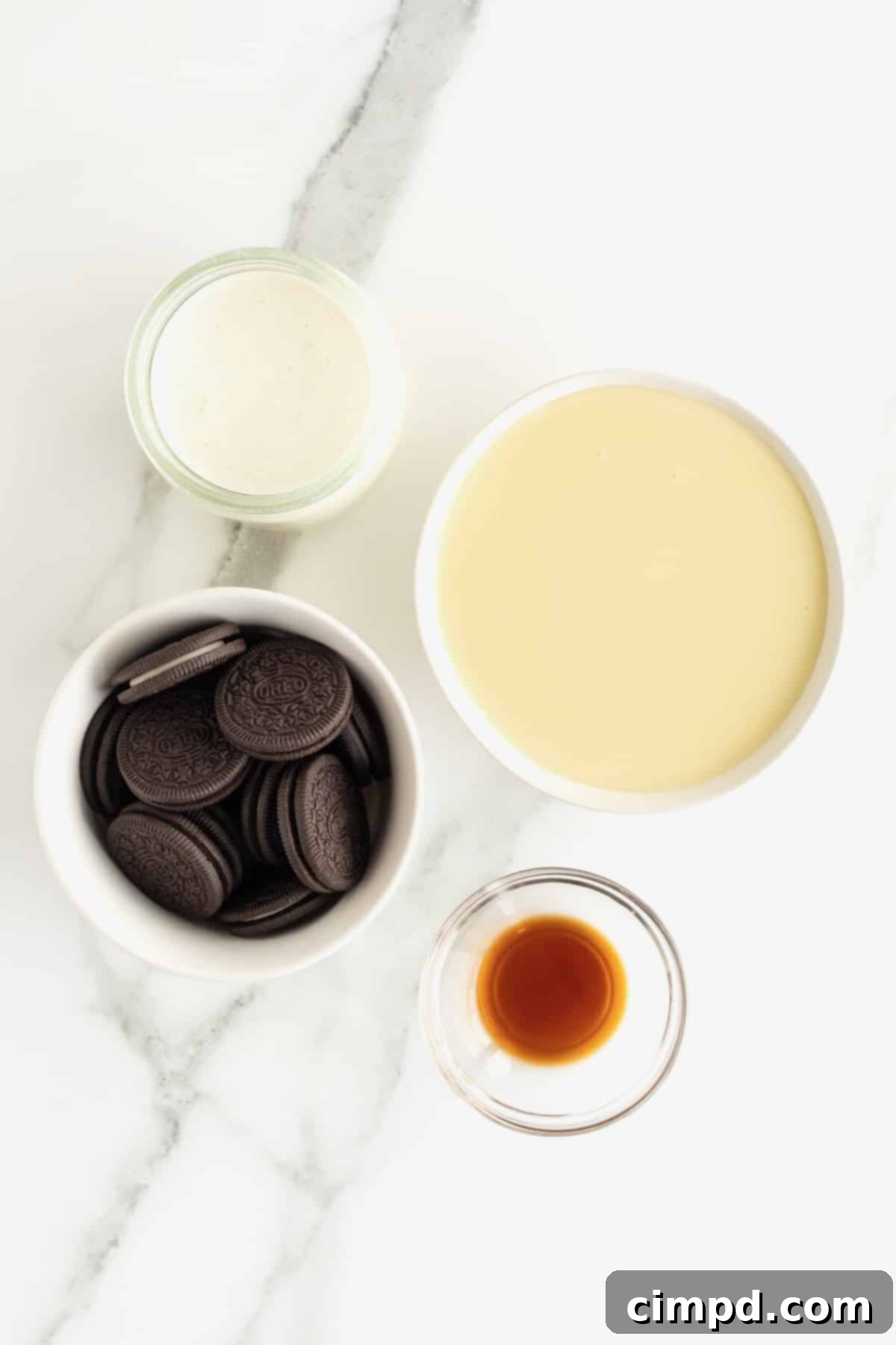 Black and White Bliss 6 A flat lay of the essential ingredients: Oreo cookies, heavy whipping cream, sweetened condensed milk, and vanilla extract, all artfully arranged in small glass bowls on a pristine white marble counter.