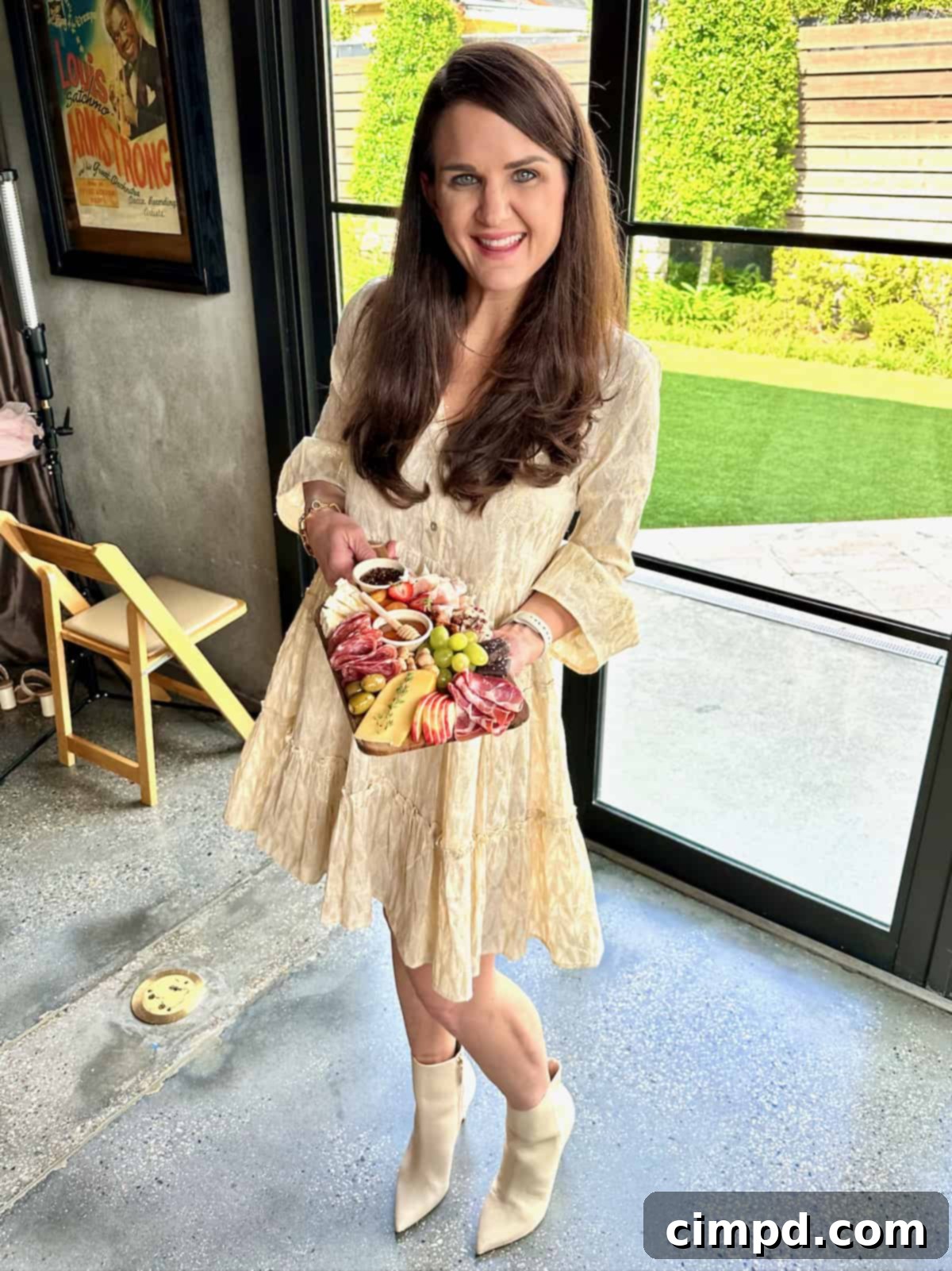 The BakerMama Maegan Brown, dressed in an elegant textured knee-length ivory dress and chic ivory pointed-toe ankle boots, holds a small, artfully arranged charcuterie board.