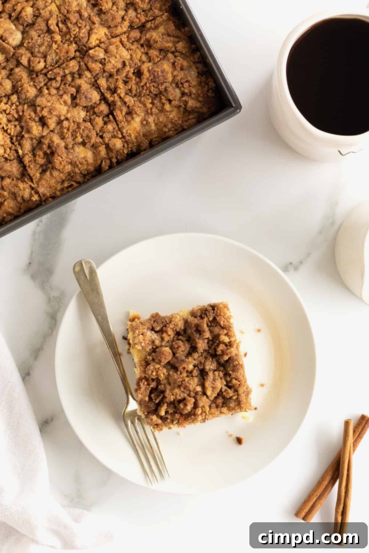 A square slice of coffee cake on a white ceramic plate with a metal fork, with a full pan of coffee cake blurred in the background.