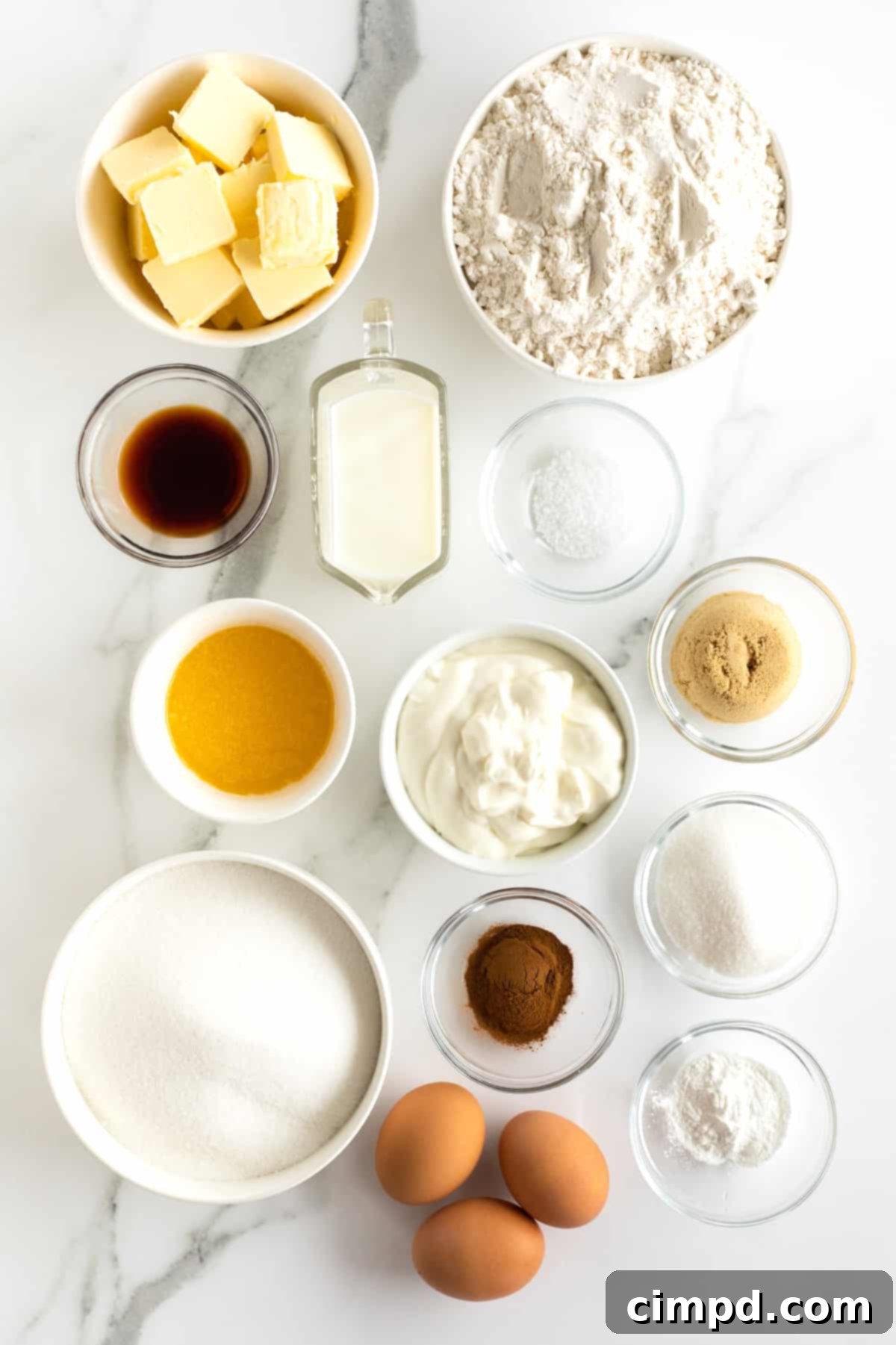 Various baking ingredients including butter, flour, milk, vanilla extract, brown sugar, granulated sugar, ground cinnamon, kosher salt, and eggs, arranged in small glass containers on a white marble counter.