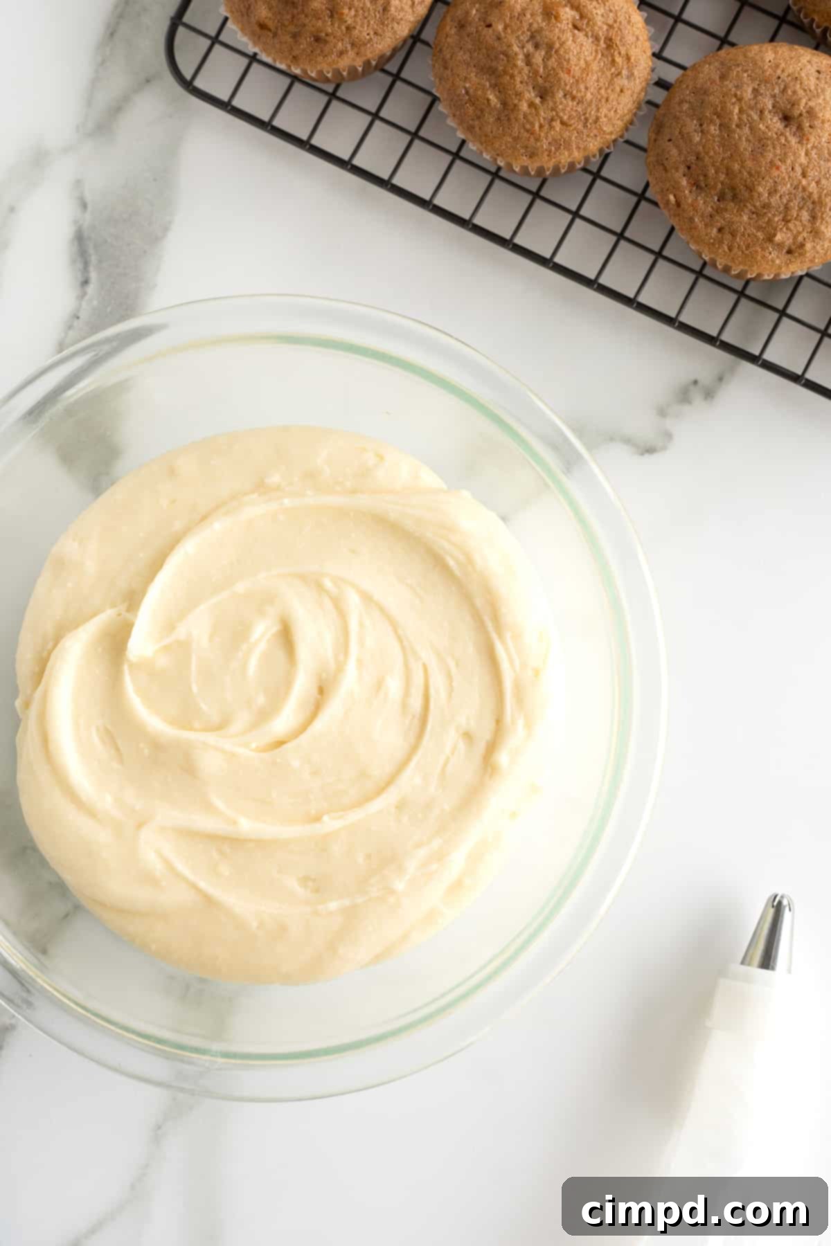 Whip Up Perfect Cream Cheese Frosting 4 A clear glass bowl of cream cheese icing sitting next to a cooling rack of cupcakes.