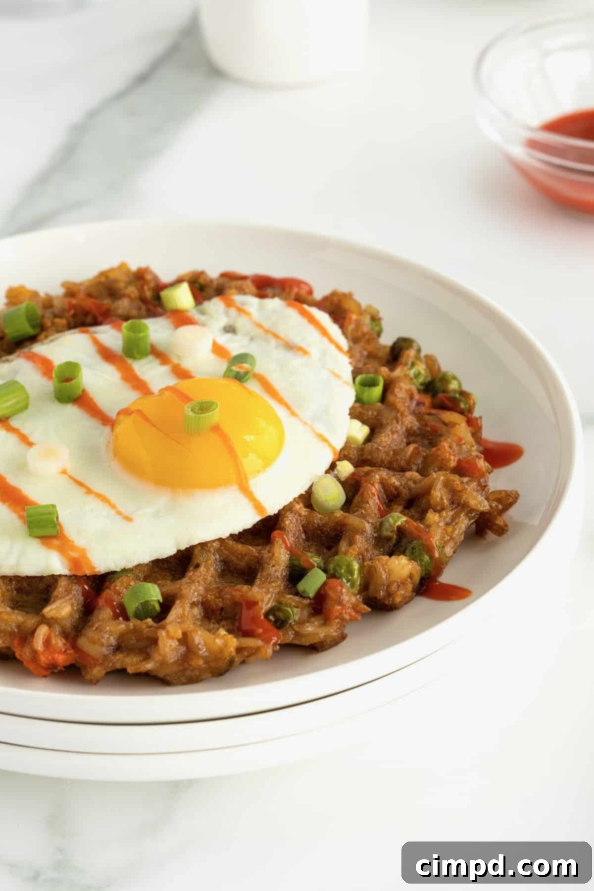 A sunny side up egg on top of a fried rice waffle on a white ceramic plate sprinkled with chopped green onion and drizzled with sriracha sauce, ready to be enjoyed.