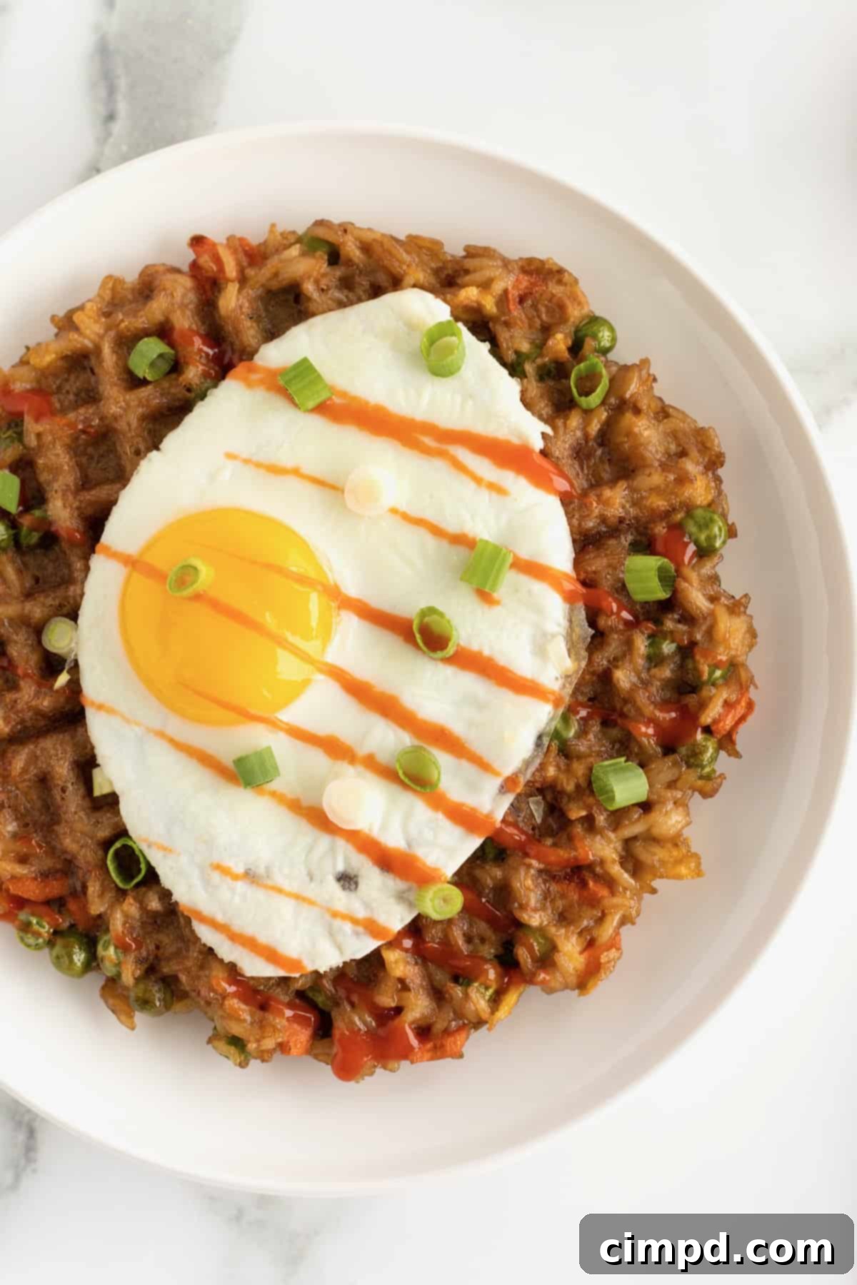 A sunny side up egg on top of a fried rice waffle on a white ceramic plate, beautifully garnished with chopped green onion and a drizzle of sriracha sauce, ready for indulgence.