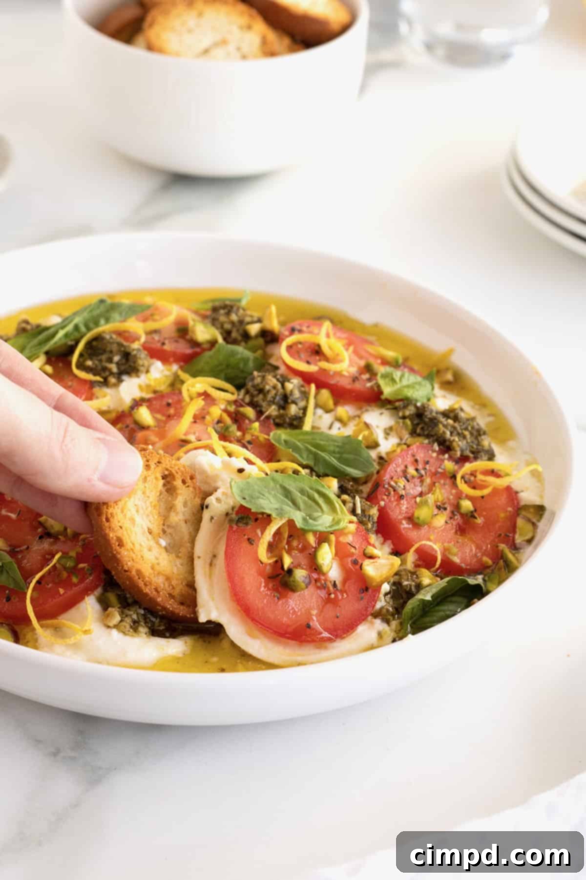 Velvet Burrata Caprese Board 3 Hand dipping crostini into burrata caprese in a white serving bowl, highlighting the creamy texture.