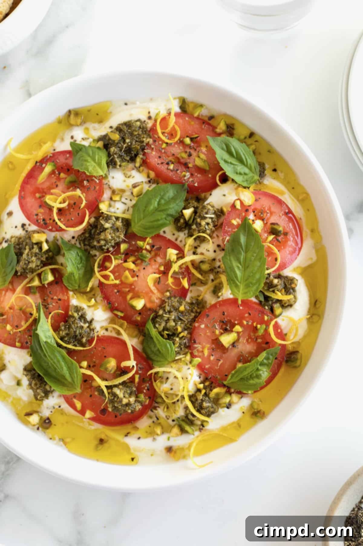 Velvet Burrata Caprese Board 5 A round white dish with sliced tomatoes, fresh basil and olive oil on top of burrata, ready to be served as a salad or platter.