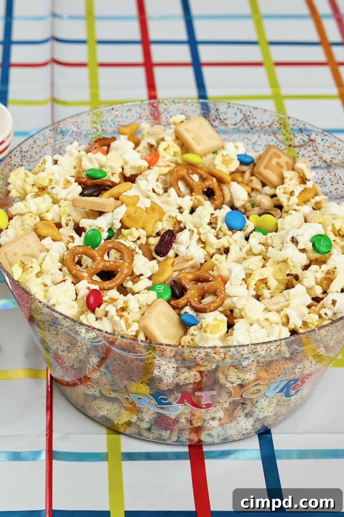 A large glass bowl reading TREAT YOURSELF filled with a popcorn snack mix with pretzels, M&Ms, dried cranberries, roasted peanuts and alphabet cookies.