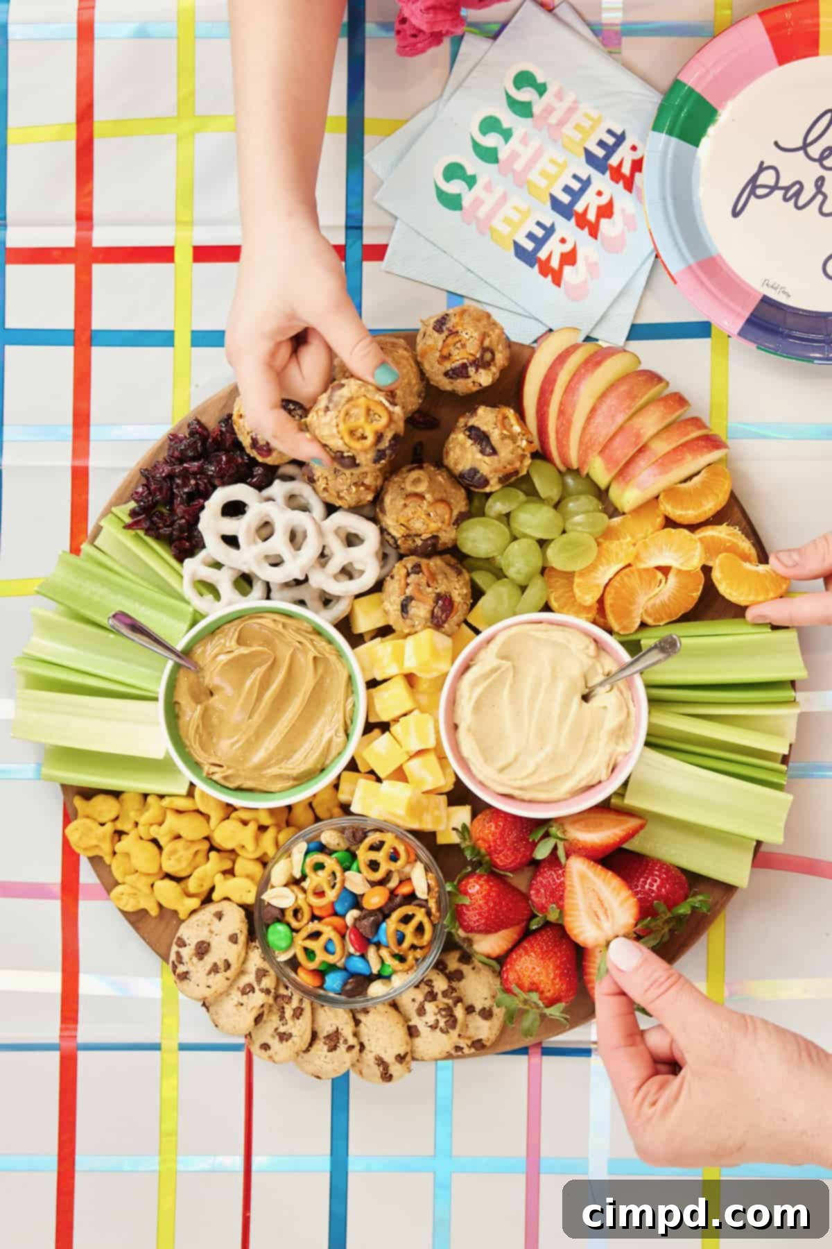 A beautifully arranged round wooden board overflowing with healthy and fun after-school snacks. It features creamy hummus and peanut butter in small bowls, alongside a homemade snack mix of peanuts, mini pretzels, chocolate chips, and M&M’s. Cubes of cheese, crisp celery sticks, fresh apple slices, bright mandarin orange segments, halved green grapes, and juicy strawberry halves add color and essential nutrients. Completing the board are crunchy Goldfish crackers, sweet dried cranberries, delicate yogurt-covered pretzels, and miniature chocolate chip cookies, offering a balanced mix of textures and tastes for hungry children.
